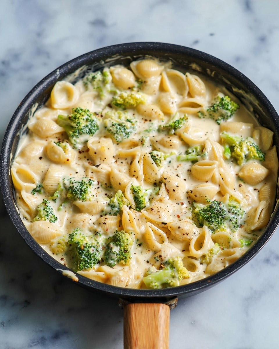 The image shows a close-up of a creamy pasta dish with small shell pasta and bright green broccoli pieces mixed together. The pasta is covered in a smooth, pale yellow sauce with a soft, slightly thick texture. Small black pepper bits are sprinkled all over the dish, adding contrast to the light colors. The broccoli florets are fresh and have a slightly rough texture, while the pasta shells are soft and curved, sitting evenly throughout. The whole dish fills a white bowl placed on a white marbled surface. Photo taken with an iphone --ar 4:5 --v 7