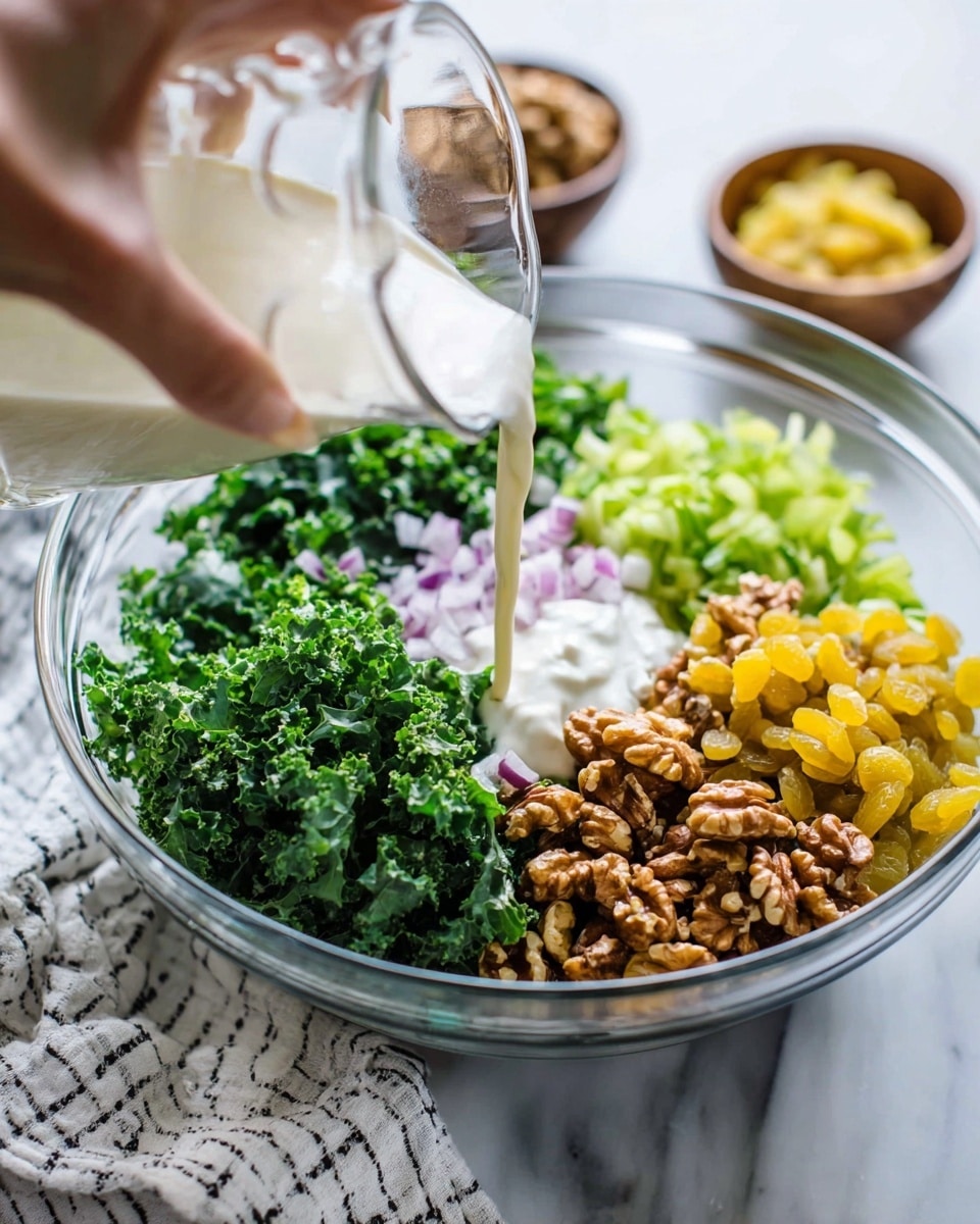 A clear glass bowl holds a fresh salad arranged in visible piles: finely chopped dark green kale on the left, shredded bright green celery next to it, yellow golden raisins at the front, and medium brown walnut halves on the right. In the center, small pieces of light purple chopped onion are scattered atop a dollop of white creamy dressing. A woman's hand is pouring more creamy dressing from a clear glass pitcher over the salad. The bowl sits on a white marbled surface with a white and black striped cloth partially under it, and blurred bowls with more ingredients in the background. Photo taken with an iphone --ar 4:5 --v 7