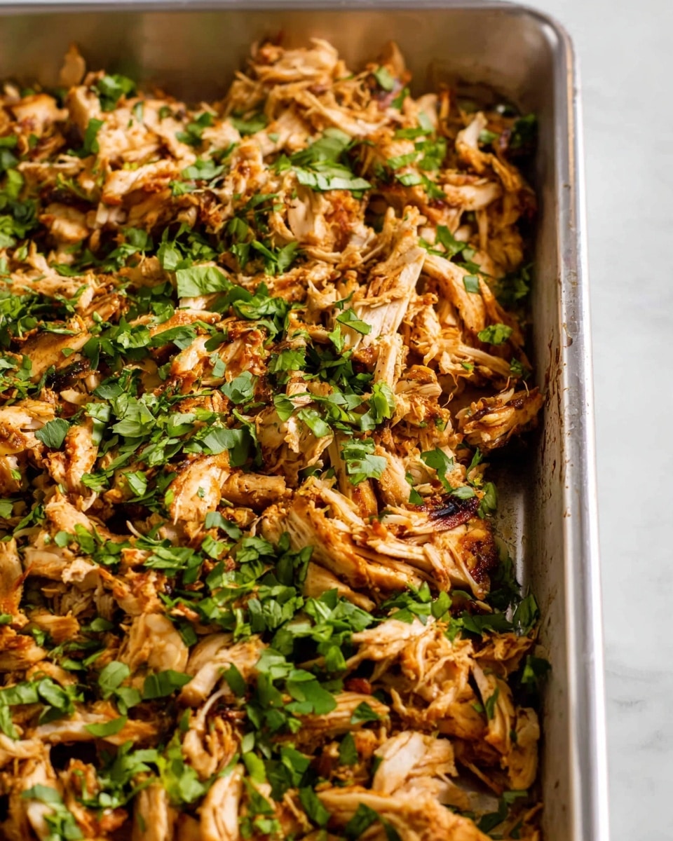 A close-up view of a metal baking tray filled with shredded, cooked chicken that has a golden-brown color with some darker grilled spots, covered evenly with chopped fresh green herbs across the top. The tray sits on a white marbled surface, and no other items are visible around it. photo taken with an iphone --ar 4:5 --v 7