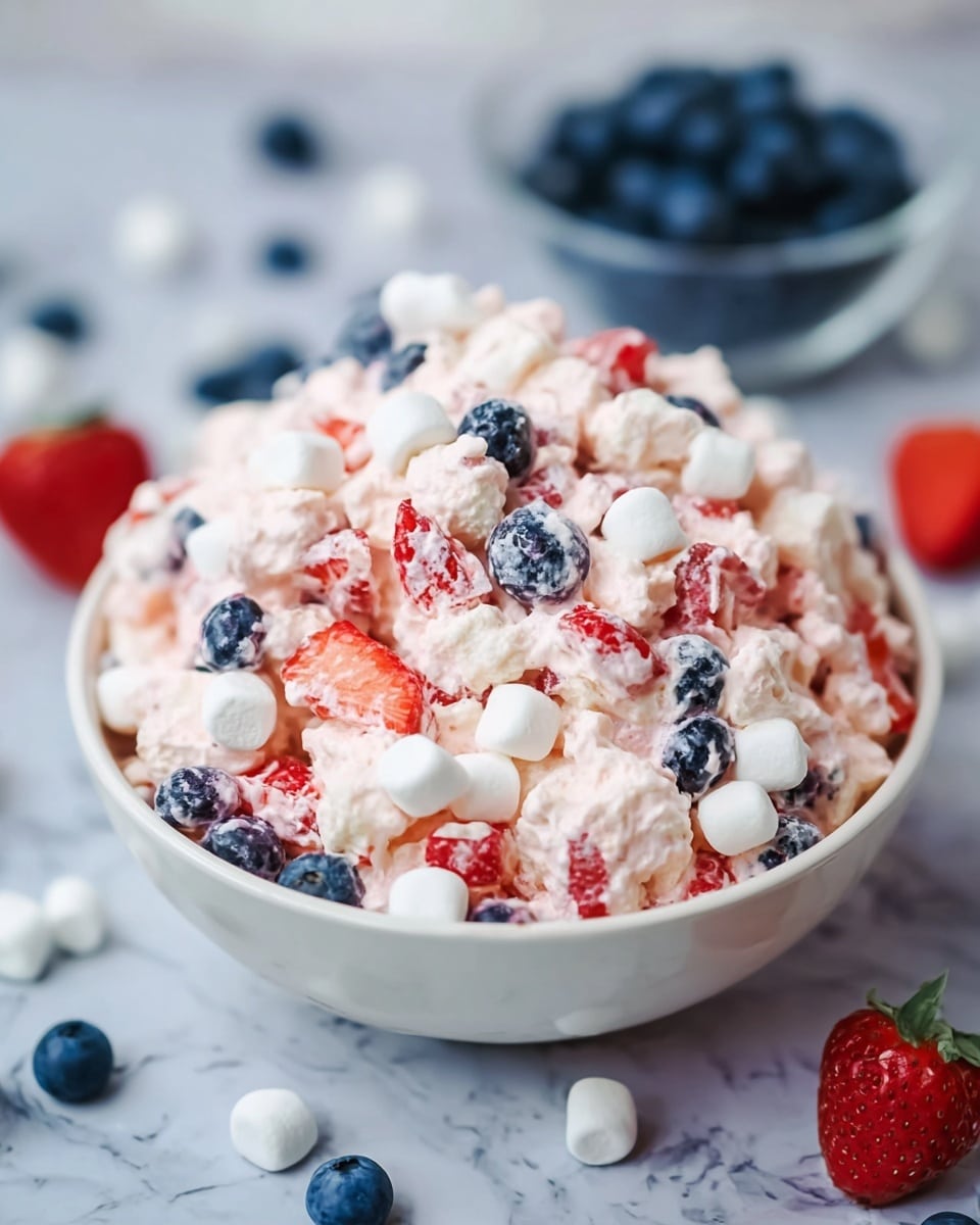 A large white bowl filled with a creamy, chunky mix of small white marshmallows, bright red sliced strawberries, and dark blue blueberries, all coated in a pale pink whipped mixture with a soft, fluffy texture. The bowl is placed on a white marbled surface with scattered blueberries, marshmallows, and a half strawberry around it. A small bowl of fresh blueberries is blurred in the background. photo taken with an iphone --ar 4:5 --v 7