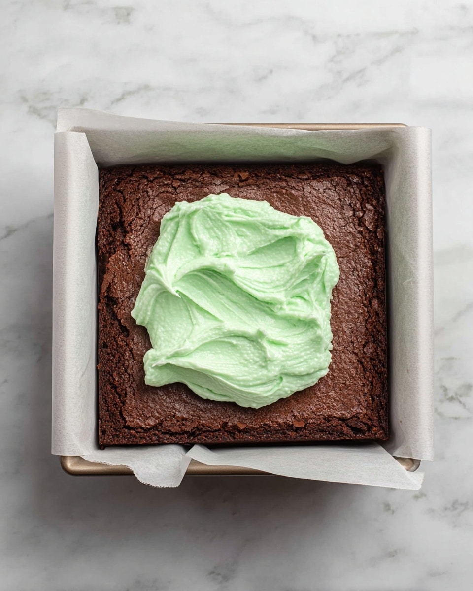 A single layer of dark brown baked brownie sits in a square baking pan lined with white parchment paper, topped in the center with a dollop of light green creamy frosting that has a soft, swirled texture. The pan rests on a white marbled surface. Photo taken with an iphone --ar 4:5 --v 7