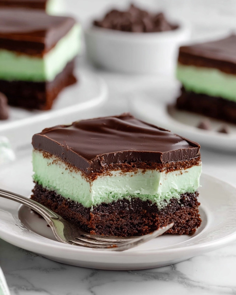This image shows a square piece of layered dessert on a white plate with a silver fork holding a small bite next to it. The dessert has three clear layers: the bottom layer is a dark, moist chocolate cake with a crumbly texture; the middle layer is a thick, smooth mint green filling; the top layer is a glossy, dark chocolate ganache with a smooth surface and slight ridges where a bite was taken from. The plate sits on a white marbled surface, with blurred background elements including more pieces of the dessert and a white bowl of chocolate chips. Photo taken with an iphone --ar 4:5 --v 7