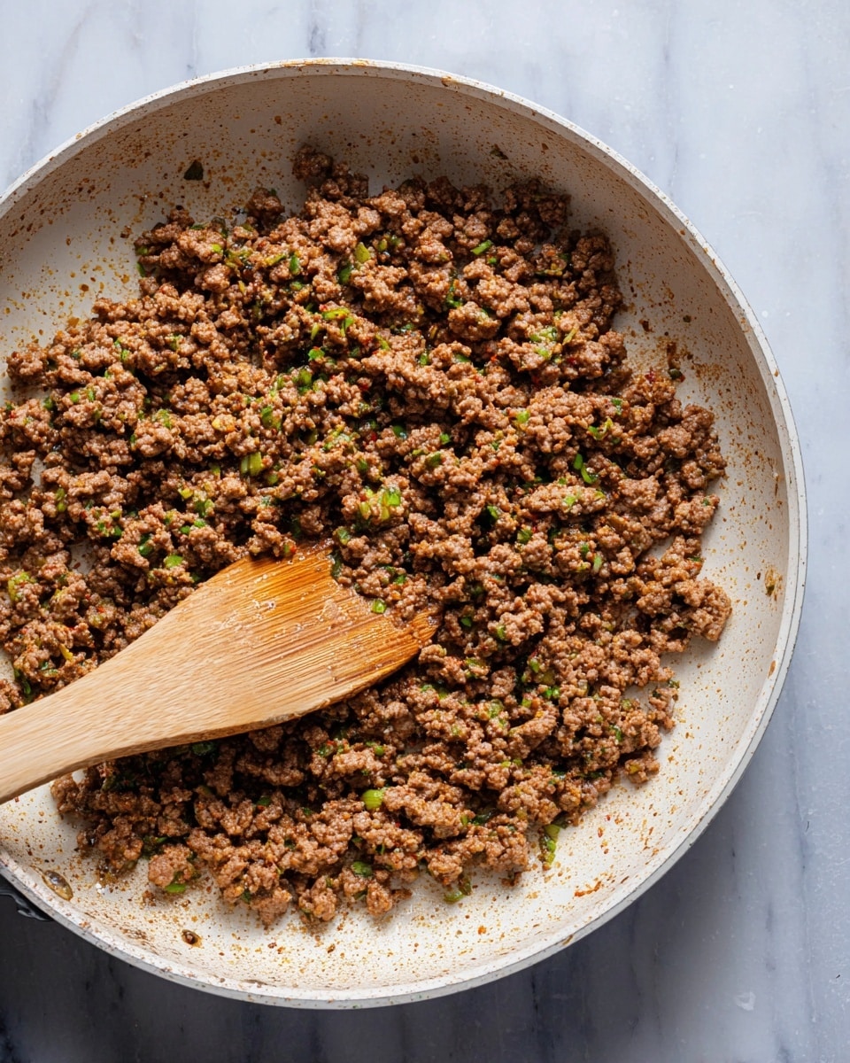 A close-up view of cooked ground meat mixed with small green herbs and spices, all inside a white pan. The meat is brown and crumbly with bits of green scattered throughout. A wooden spoon is resting inside the pan, stirring some of the meat, showing a smooth texture on part of it. The pan sits on a white marbled surface with no other items visible. photo taken with an iphone --ar 4:5 --v 7