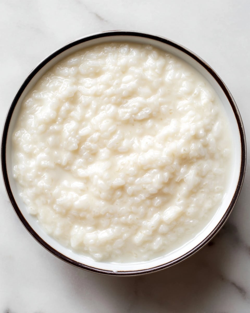 A close-up top view of a bowl filled with creamy rice pudding that has a soft, smooth texture with visible individual white rice grains mixed evenly throughout the thick, milky base. The single layer of pudding is creamy white with a slightly shiny surface, all contained in a white bowl with a thin dark rim, placed on a white marbled texture background. photo taken with an iphone --ar 4:5 --v 7