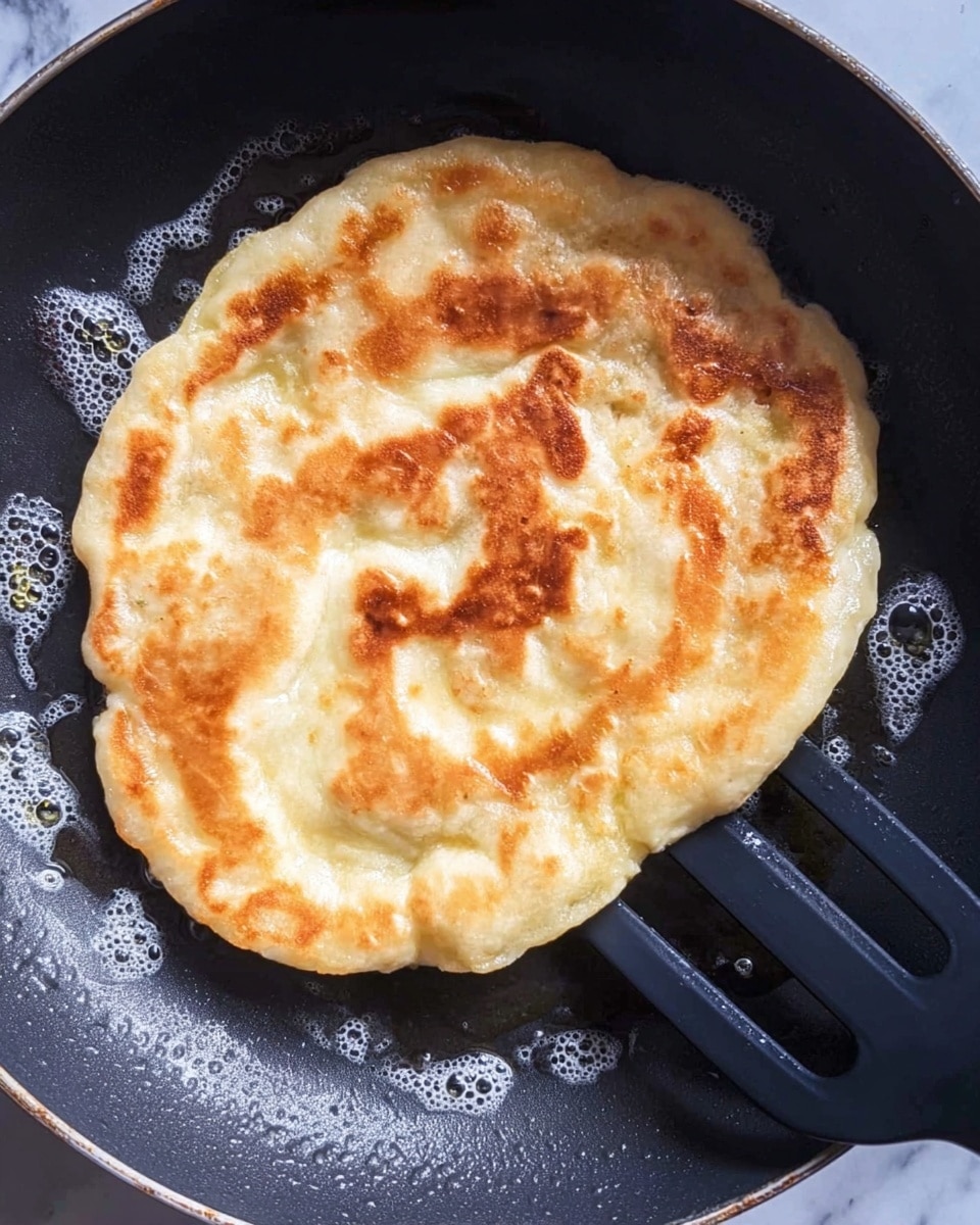 A single flat round piece of dough is frying in a black pan with bubbles and a golden brown texture on the top surface. The dough is puffed up in some areas, showing a soft and uneven texture, with light yellow and white tones underneath the golden brown spots. A black spatula is partially under the dough on the right side, ready to lift it. The pan has small drops of oil glistening around the dough. The background is a white marbled texture. photo taken with an iphone --ar 4:5 --v 7