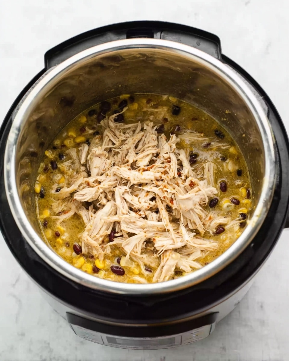 The image shows a close-up of an open Instant Pot filled with shredded light brown chicken sitting on top of a yellow broth mixed with small black beans and diced yellow vegetables like corn. The metal inner pot has a shiny silver surface with some bits of food on the sides. The Instant Pot itself has a black and silver exterior with a control panel visible at the front. The whole scene is set on a white marbled surface. photo taken with an iphone --ar 4:5 --v 7