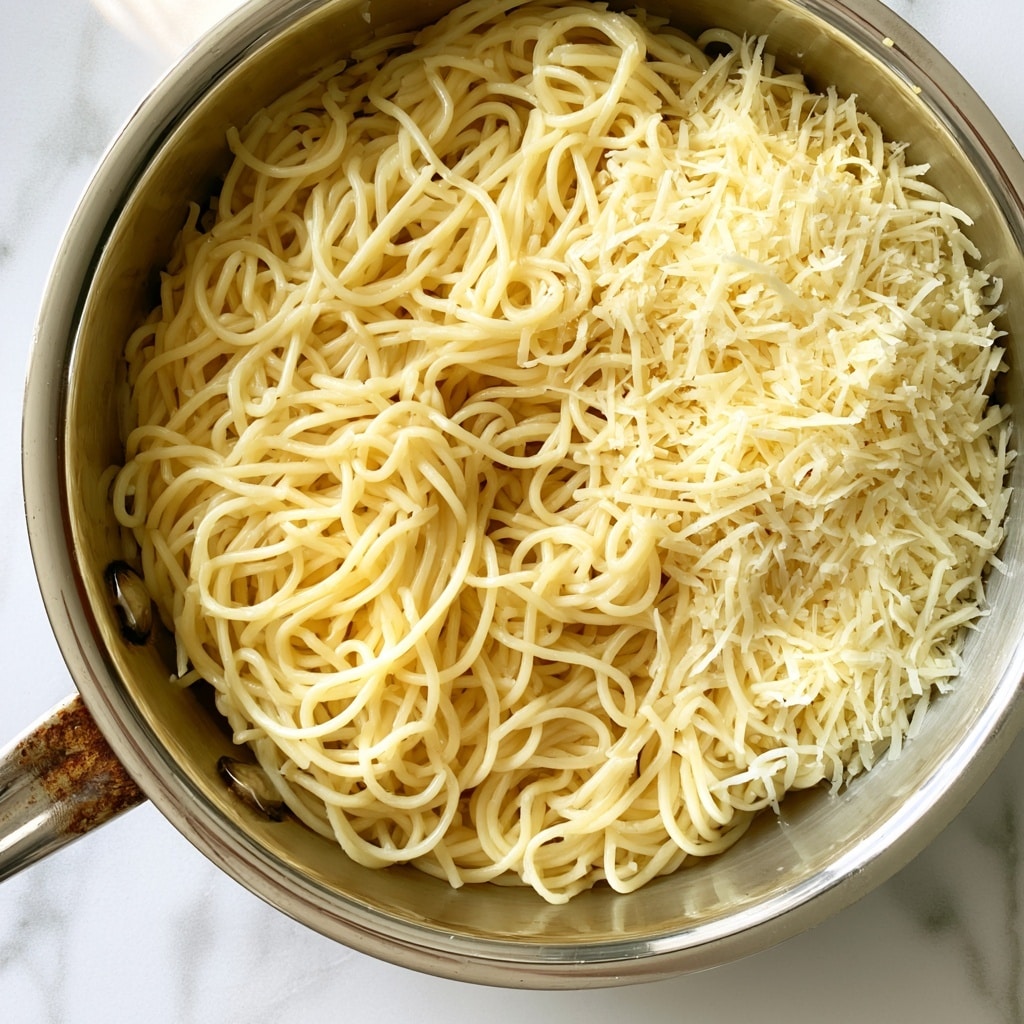 A shiny silver pan is filled with light yellow cooked spaghetti that looks soft and slightly glossy. On top of the pasta, there is a loose layer of finely shredded pale yellow cheese mostly gathered on one side. The pan is placed on a clean white marbled surface with soft gray veins. The metal handle of the pan is visible on the left side, showing some wear and rust stains. The entire scene has soft natural light coming from above, highlighting the texture of the noodles and cheese. photo taken with an iphone --ar 4:5 --v 7