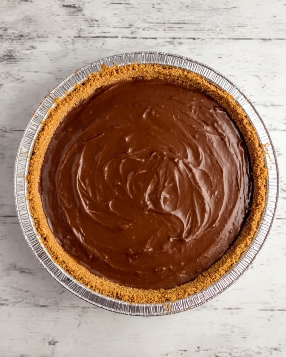 The image shows a pie with three clear layers. The bottom crust is golden brown and crumbly, holding the filling inside a round foil pie dish. Above the crust is a thick, smooth chocolate filling that is shiny and dark brown, filling the pie up to the edges of the crust. The surface of the filling has light swirling patterns, adding texture to the smooth chocolate top. The pie sits on a white marbled surface. Photo taken with an iphone --ar 4:5 --v 7