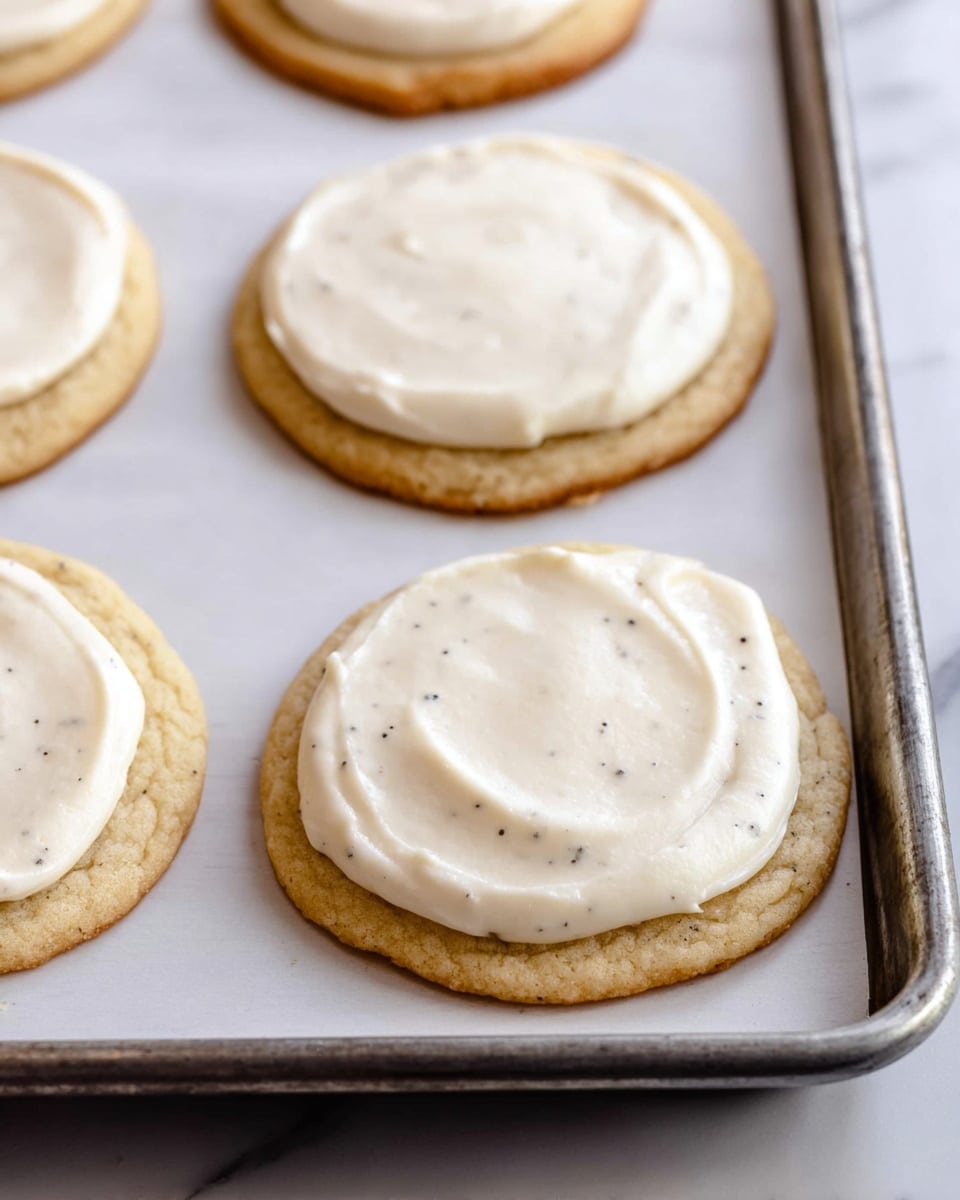 The image shows several round cookies on a baking sheet lined with white parchment paper, each cookie has one thick layer of smooth, creamy white frosting spread evenly on top, with tiny dark specks visible in the frosting. The cookies are golden brown with a soft texture visible around the edges, placed close to each other in rows on the metal tray, which rests on a white marbled surface. photo taken with an iphone --ar 4:5 --v 7