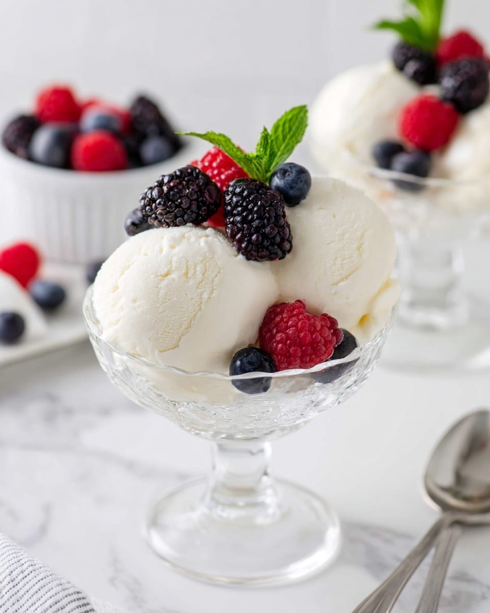 The image shows two smooth, round scoops of white ice cream served in a clear glass bowl on a white marbled surface. Around the ice cream, there are small clusters of dark purple blackberries and bright blue blueberries. On top of the scoops, a few fresh green mint leaves add a pop of color along with a couple of red raspberries, giving the dish a fresh and colorful look. photo taken with an iphone --ar 4:5 --v 7