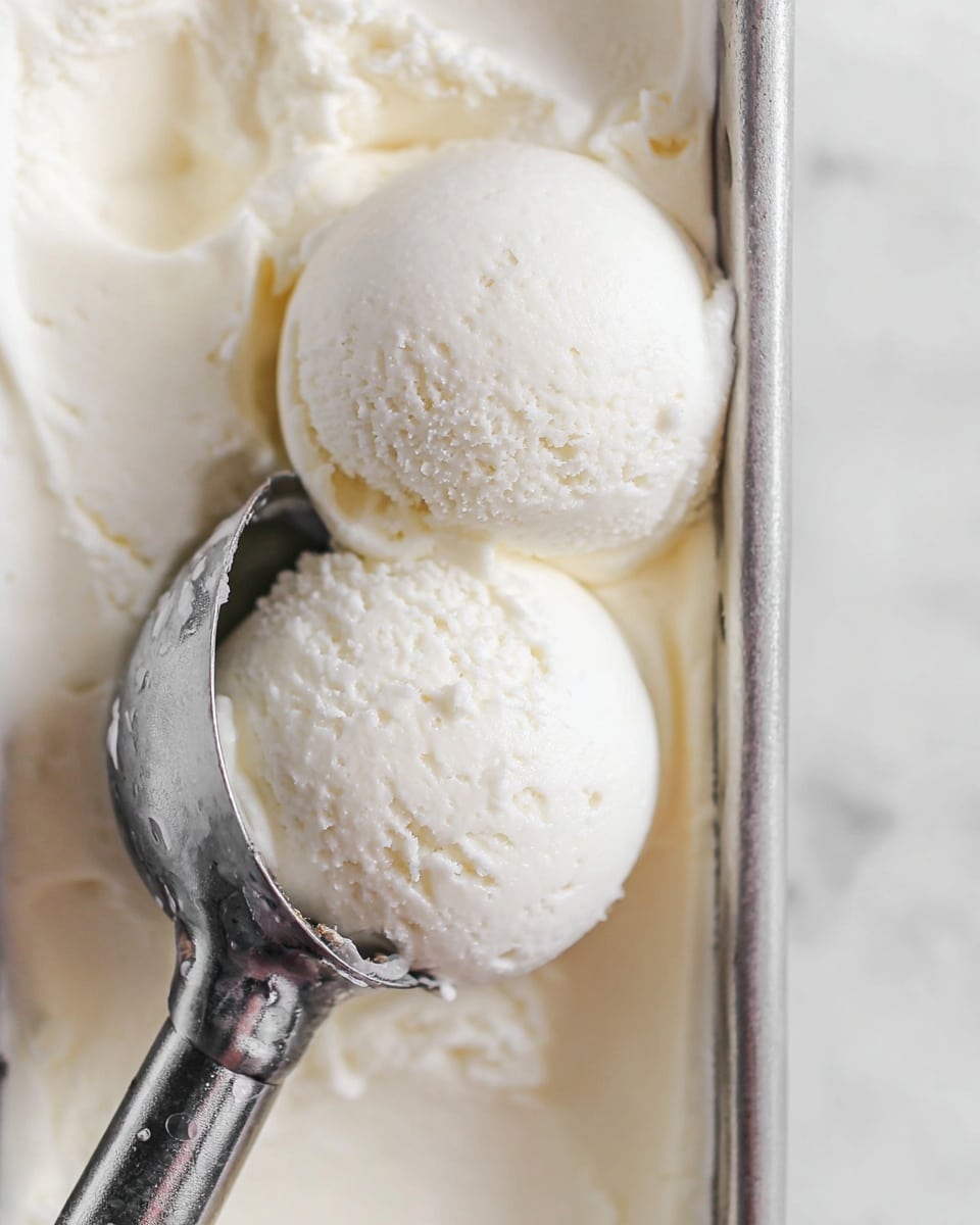 A close-up view of two smooth, round scoops of creamy white ice cream held by a metal ice cream scooper resting inside a silver container filled with more white ice cream, all placed on a white marbled surface that softly reflects the light, emphasizing the cool texture and pure color of the ice cream. photo taken with an iphone --ar 4:5 --v 7