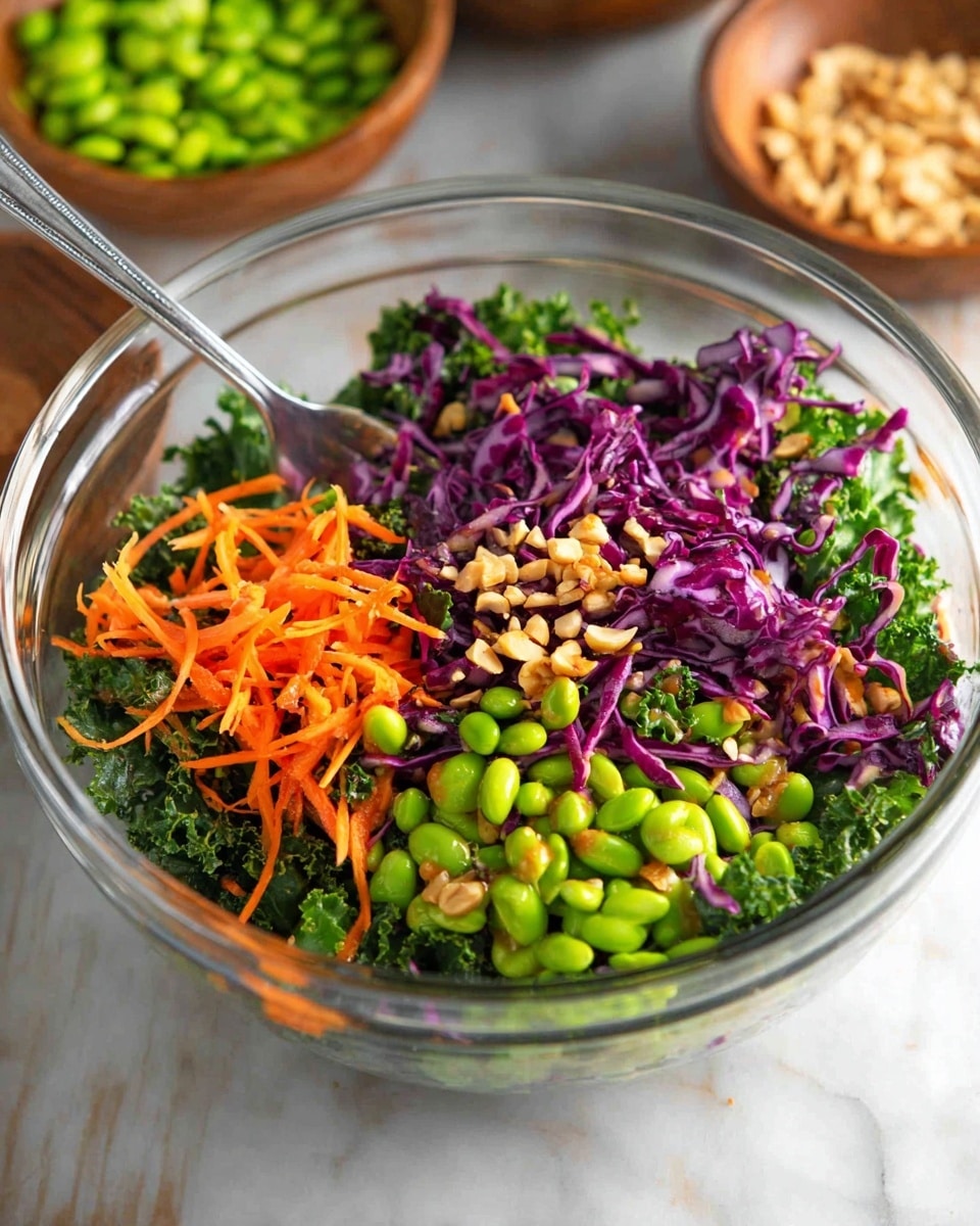 A clear glass bowl filled with a colorful salad sits on a wooden surface, featuring three main layers: a base of dark green kale leaves with a rough texture, topped with thin strips of bright orange carrot and purple cabbage scattered throughout, and a generous layer of bright green edamame beans. Small pieces of chopped almonds are sprinkled on top for texture. A silver spoon rests inside the bowl, and in the background, two wooden bowls hold extra edamame beans and chopped almonds. The whole scene is set against a white marbled texture. photo taken with an iphone --ar 4:5 --v 7