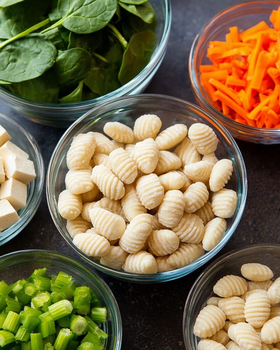 A clear glass bowl filled with small oval-shaped gnocchi, pale off-white colored with visible ridges, is placed in the front center. Behind this bowl, there are four more clear glass bowls arranged in a loose circle: one with bright green fresh spinach leaves on the left, one with small cubes of pale white tofu in the top left, one with thin orange carrot sticks to the top right, and one with bright green chopped celery slices in the top right. The bowls sit on a dark surface that contrasts with the clear glass bowls and colorful ingredients. photo taken with an iphone --ar 4:5 --v 7