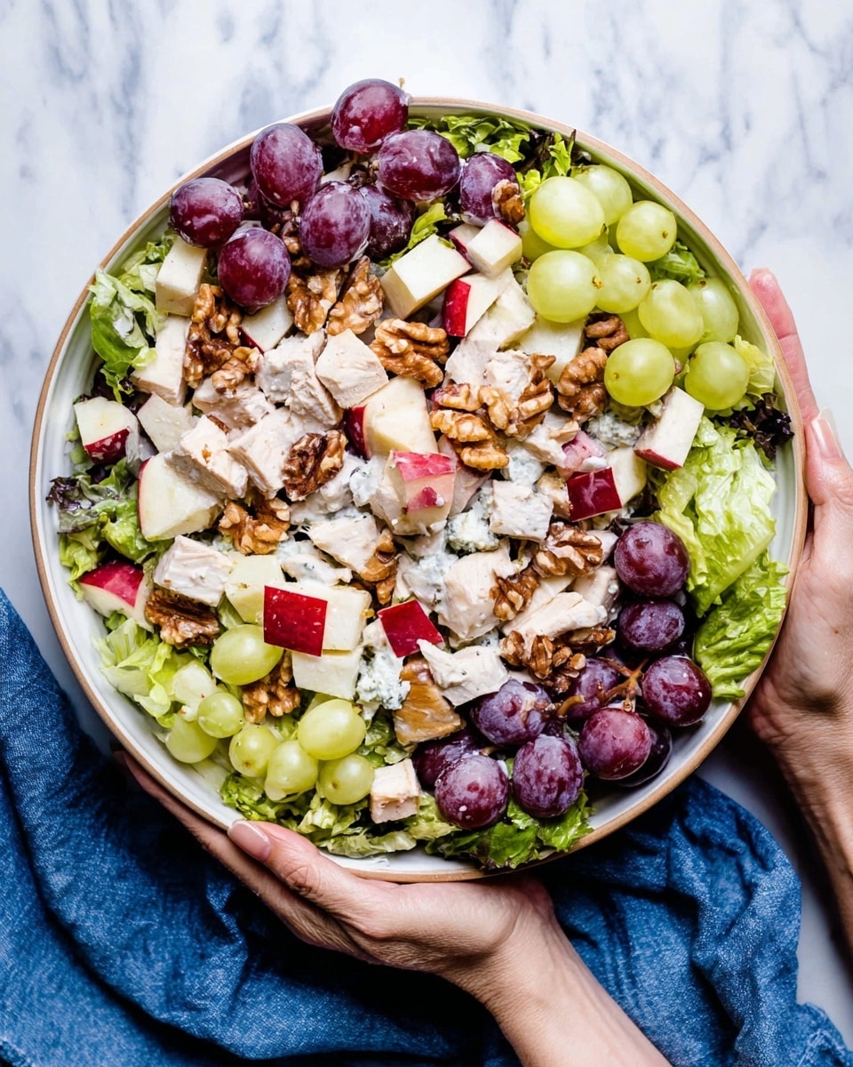 A large bowl full of fresh salad with layers starting with green and purple lettuce leaves at the bottom, topped with creamy white chunks of chicken spread evenly, bright red apple pieces, halved purple grapes, and pale green celery slices. Scattered over the top are walnut halves to add texture. The bowl is white and is held gently by two women's hands on a white marbled surface with a blue cloth nearby. photo taken with an iphone --ar 4:5 --v 7