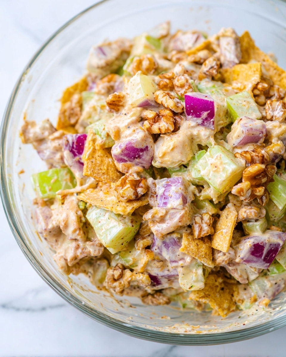 A close-up view of a creamy salad mixed in a clear glass bowl on a white marbled surface, showing layers of pale yellow tortilla chip pieces, light green celery chunks, bright purple-red onion pieces, and walnuts coated in a thick, light beige creamy dressing with visible pepper and spice flecks. The salad has a chunky texture with varied shapes and colors mixed together, highlighted by the shiny and smooth cream sauce covering all ingredients photo taken with an iphone --ar 4:5 --v 7