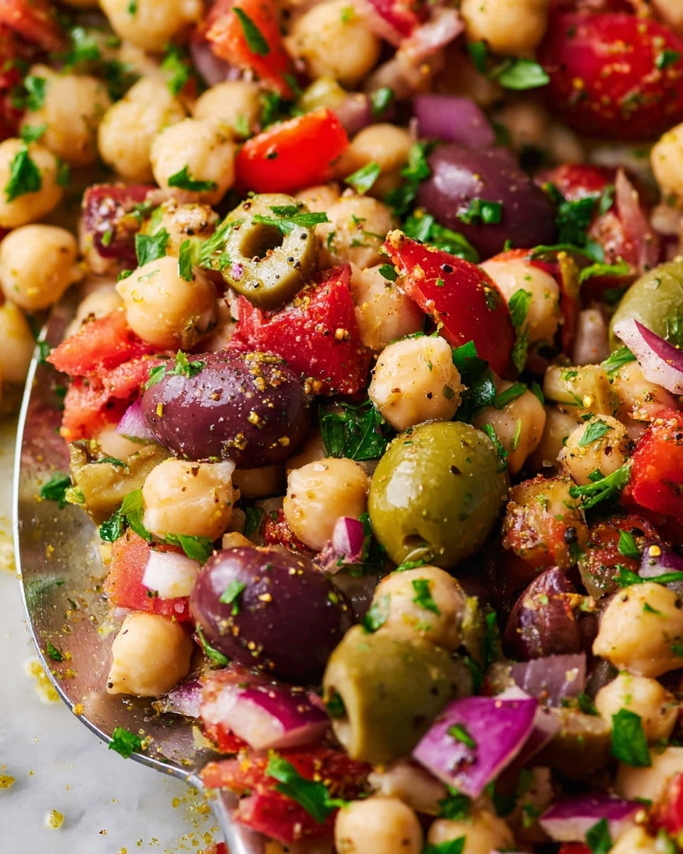 A close-up view of a colorful chickpea salad with three main layers evenly mixed throughout: round, light beige chickpeas, green and dark purple olives, and bright chopped vegetables including red tomatoes and red onions. Fresh green herbs are scattered on top, and a sprinkle of seasoning adds texture on the olives and chickpeas. All ingredients appear fresh, juicy, and shiny, resting on a white marbled surface with a clear serving spoon partially visible underneath. photo taken with an iphone --ar 4:5 --v 7