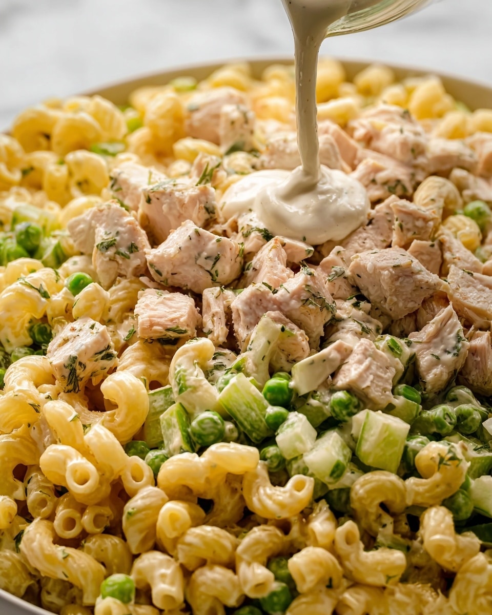 This close-up image shows a creamy pasta salad with three visible layers: the bottom layer is curved yellow pasta tubes with a smooth texture, mixed with small pieces of green peas and celery slices; the middle layer has light tan chunks of chicken coated in herbs, scattered evenly throughout; the top layer features a thick, creamy white sauce being poured over the pasta and chicken. The whole dish sits on a white bowl placed on a white marbled surface. photo taken with an iphone --ar 4:5 --v 7
