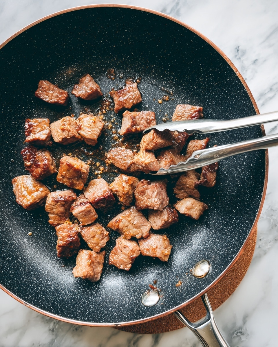 A close-up view of small, browned meat cubes cooking in a black non-stick frying pan with a speckled texture. The meat pieces are light brown with a slightly crispy surface, scattered evenly across the pan. A pair of silver metal tongs grips one piece of meat in the upper center of the pan. The pan handle, copper-colored with a silver end, points forward, resting on a brown cork pad. The background features a white marbled surface. photo taken with an iphone --ar 4:5 --v 7