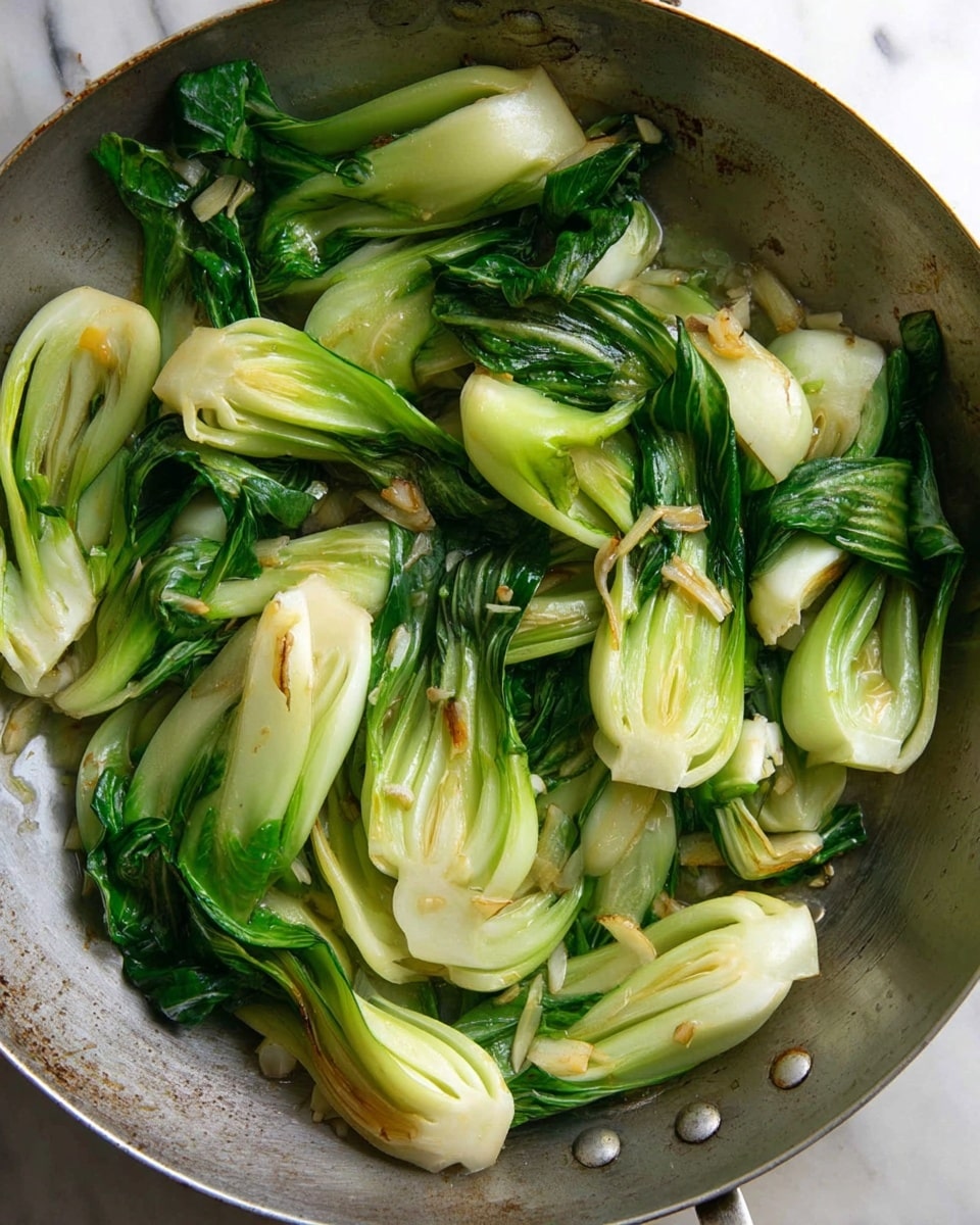 A close-up view inside a large round pan filled with cooked bok choy cut into halves or quarters showing bright green leafy tops and pale green to white thick stems. The bok choy pieces are glossy with light oil and small bits of cooked garlic and onion scattered over them, adding texture and a bit of light brown color. The pan has a worn metal look and sits on a white marbled surface. photo taken with an iphone --ar 4:5 --v 7