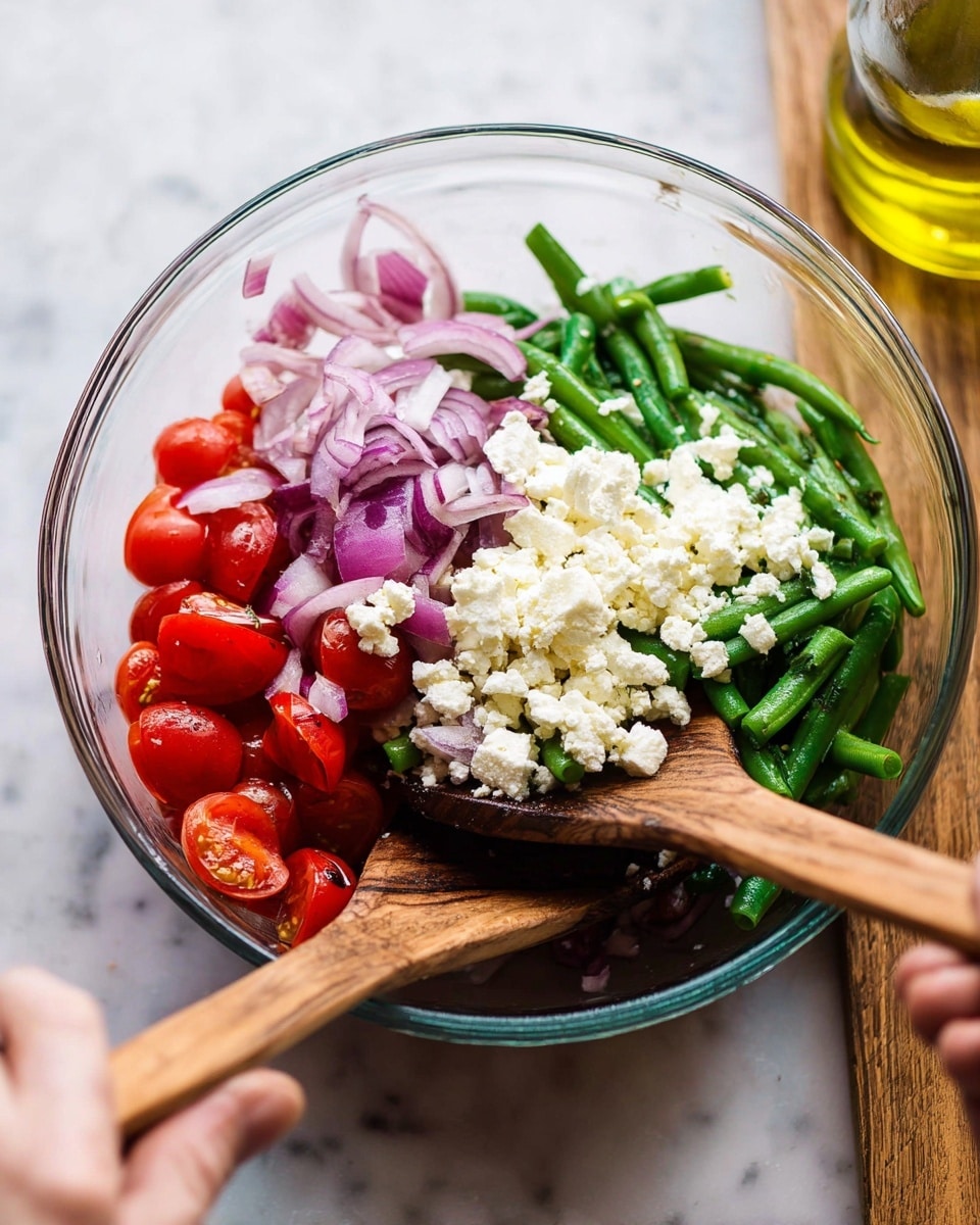 In a clear glass bowl, there is a colorful salad with four main layers: bright red cherry tomato halves at the bottom left, green beans spread mostly to the right and middle, crumbled white cheese scattered on top of the green beans, and thinly sliced purple onions placed on the top left side. There are two wooden salad utensils resting inside the bowl, one on the left and one on the right, and a woman's hand is holding the wooden spoon on the right. The bowl is placed on a white marbled surface with a glass bottle of olive oil blurred in the background. photo taken with an iphone --ar 4:5 --v 7