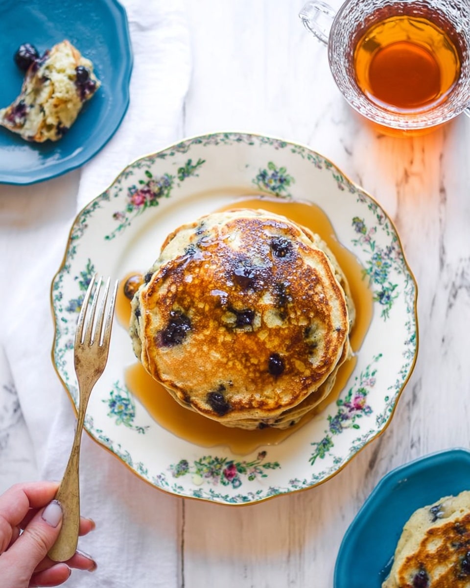 A stack of five thick pancakes filled with dark blueberries, placed on a white plate with a blue ring around the edge, sits on a white marbled surface. The top pancake is covered with shiny, dripping golden syrup that pools around the base, and the top pancake has a wedge cut out, showing the light beige inside with blueberries visible between the layers. In the background, there is a glass filled with amber liquid, slightly out of focus. photo taken with an iphone --ar 4:5 --v 7