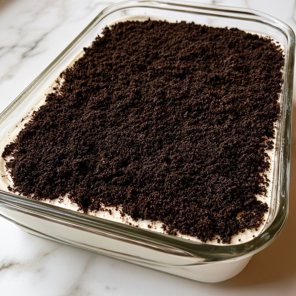 A rectangular clear glass dish holds a layered dessert with two visible layers. The bottom layer is smooth and white, covering the entire base of the dish, and appears creamy. On top is a thick, even layer of dark, finely crushed cookie crumbs, almost black, that completely cover the white layer beneath. The dish is placed on a white marbled surface, enhancing the contrast between the dark crumb layer and the light creamy layer below. The dessert has a clean and simple look, with distinct separation between the two layers, and the crumb layer has a slightly coarse texture. photo taken with an iphone --ar 4:5 --v 7