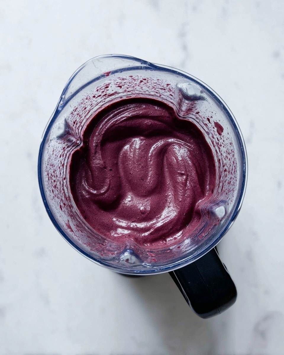 A top view of a blender with a smooth, thick dark purple mixture inside. The mixture has a creamy texture with soft swirls and a slight sheen, filling most of the blender container. The blender is placed on a white marbled surface, and its clear glass sides show some residue of the purple mix. The blender handle is black and positioned at the bottom right of the image. photo taken with an iphone --ar 4:5 --v 7