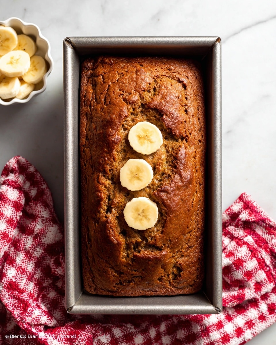 A loaf of banana bread with a rich brown, slightly cracked surface filling a rectangular metal baking pan. On top, there are three round, pale yellow banana slices arranged in the center. To the left, a small white scalloped bowl holds more banana slices, and on the right side, a red and white checkered cloth is partially visible. All items rest on a white marbled surface. photo taken with an iphone --ar 4:5 --v 7