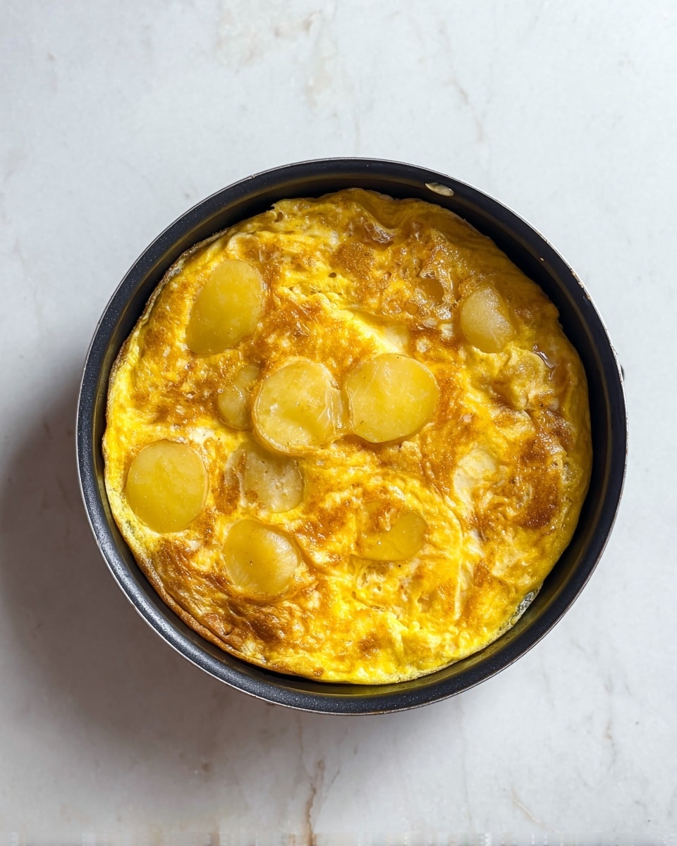 A round black pan holds a cooked omelet with visible layers of yellow egg and slices of light golden potatoes mixed in. The omelet surface is shiny and slightly bubbly with a golden yellow color and some light brown spots. The edges are soft and slightly curled in the black pan. The pan rests on a white marbled surface, creating a clean and simple look. photo taken with an iphone --ar 4:5 --v 7