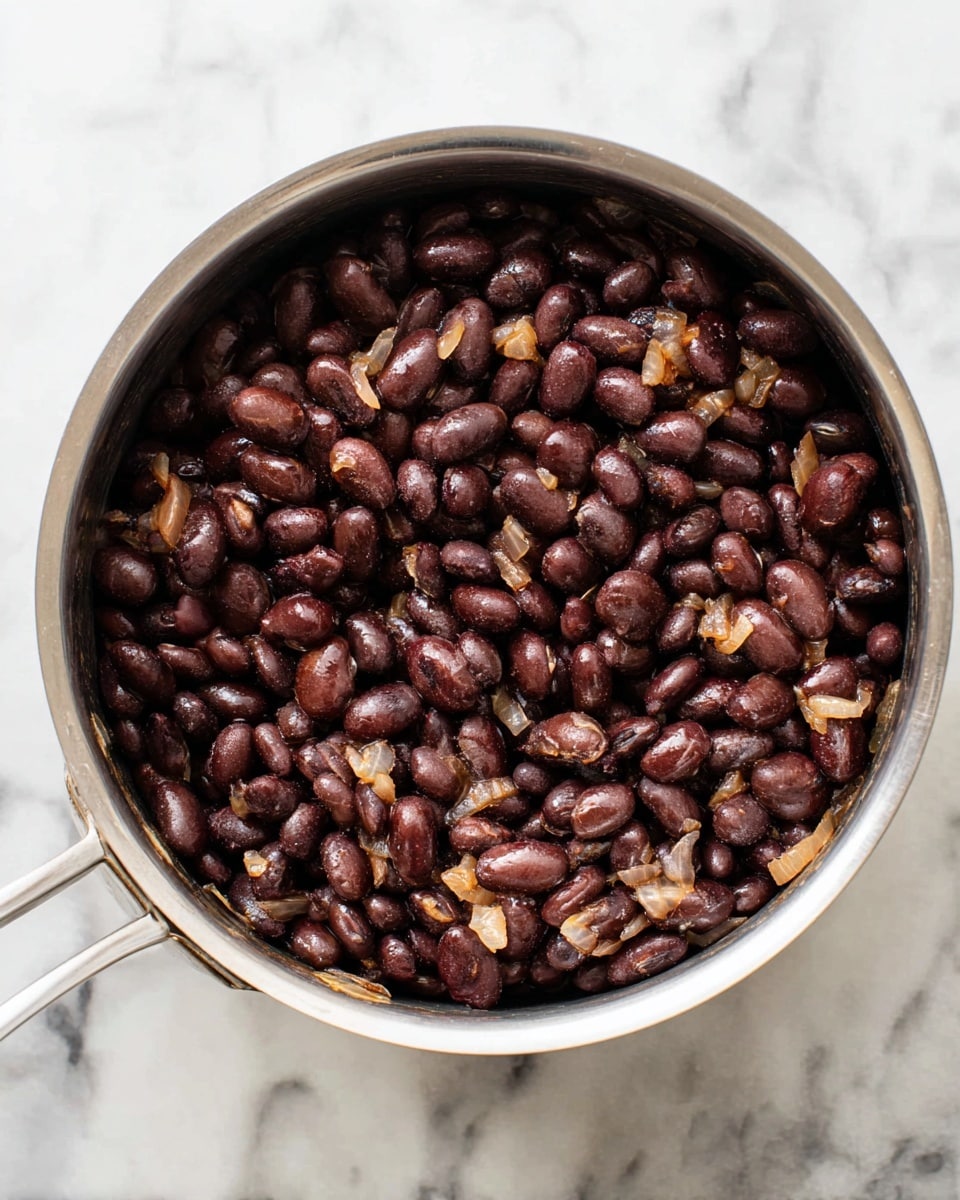 A silver pot filled with cooked black beans that have a shiny, soft texture. Mixed in are small pieces of cooked onions that are light brown and slightly translucent, scattered evenly throughout the beans. The pot is placed on a white marbled surface with subtle grey veins. The image is focused close up on the beans and onions, showing their texture clearly. Photo taken with an iphone --ar 4:5 --v 7