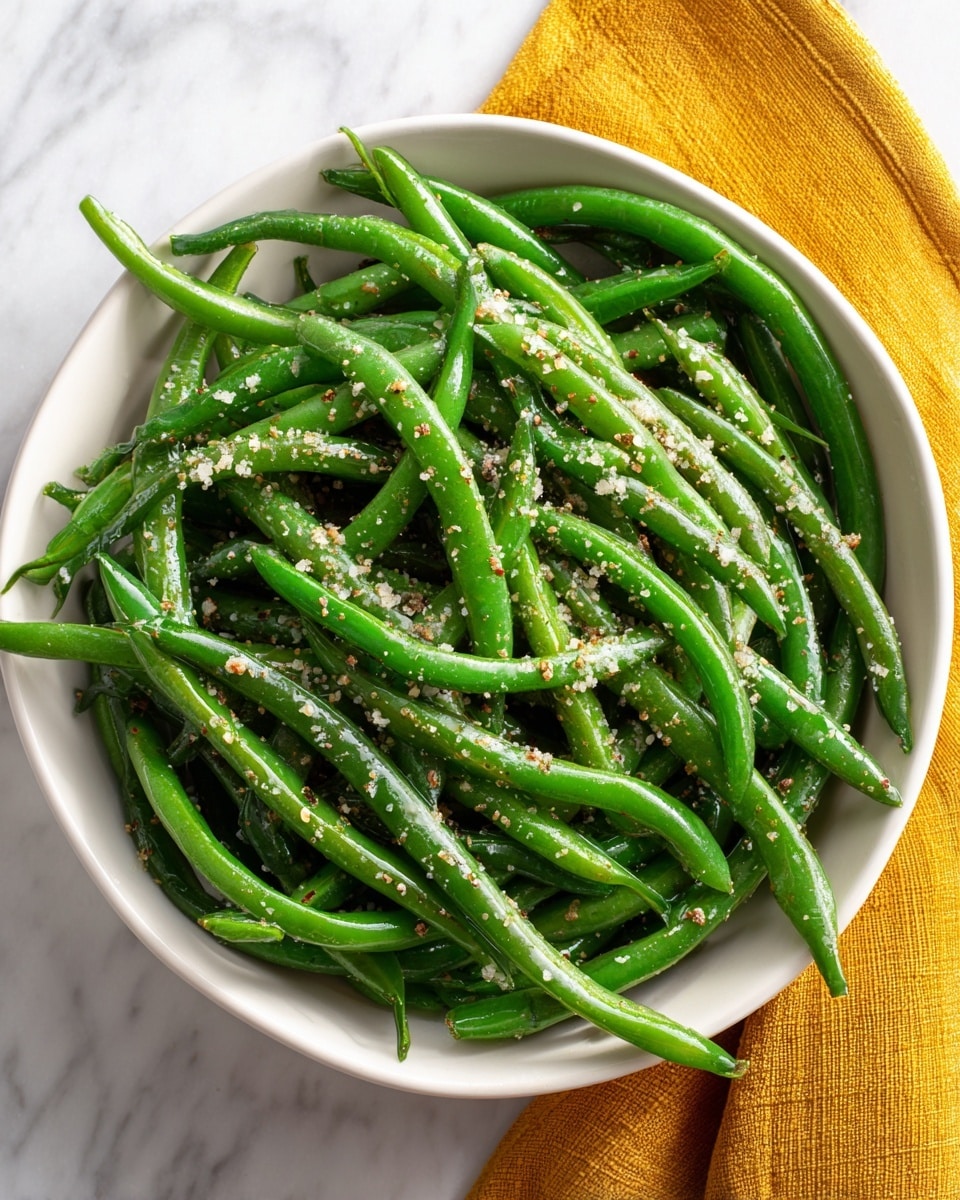 A white bowl with about one layer of fresh green beans, bright and firm, scattered evenly inside. On top of the green beans, there are small sprinkles of coarse salt, black pepper, and a light dusting of beige seasoning, adding texture and color contrast. The bowl sits on a white marbled surface with a mustard yellow cloth partially visible on the right side. photo taken with an iphone --ar 4:5 --v 7