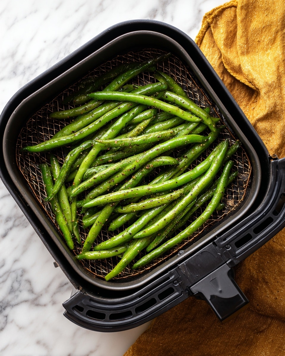 Inside a black air fryer basket with a mesh bottom, there are about twelve fresh green beans arranged in a single layer, showing their smooth and shiny texture with small black pepper specks on them. The air fryer is set on a white marbled surface with a mustard yellow cloth partially visible on the right side. The green beans are bright green and evenly spaced, highlighting their natural, firm texture. photo taken with an iphone --ar 4:5 --v 7