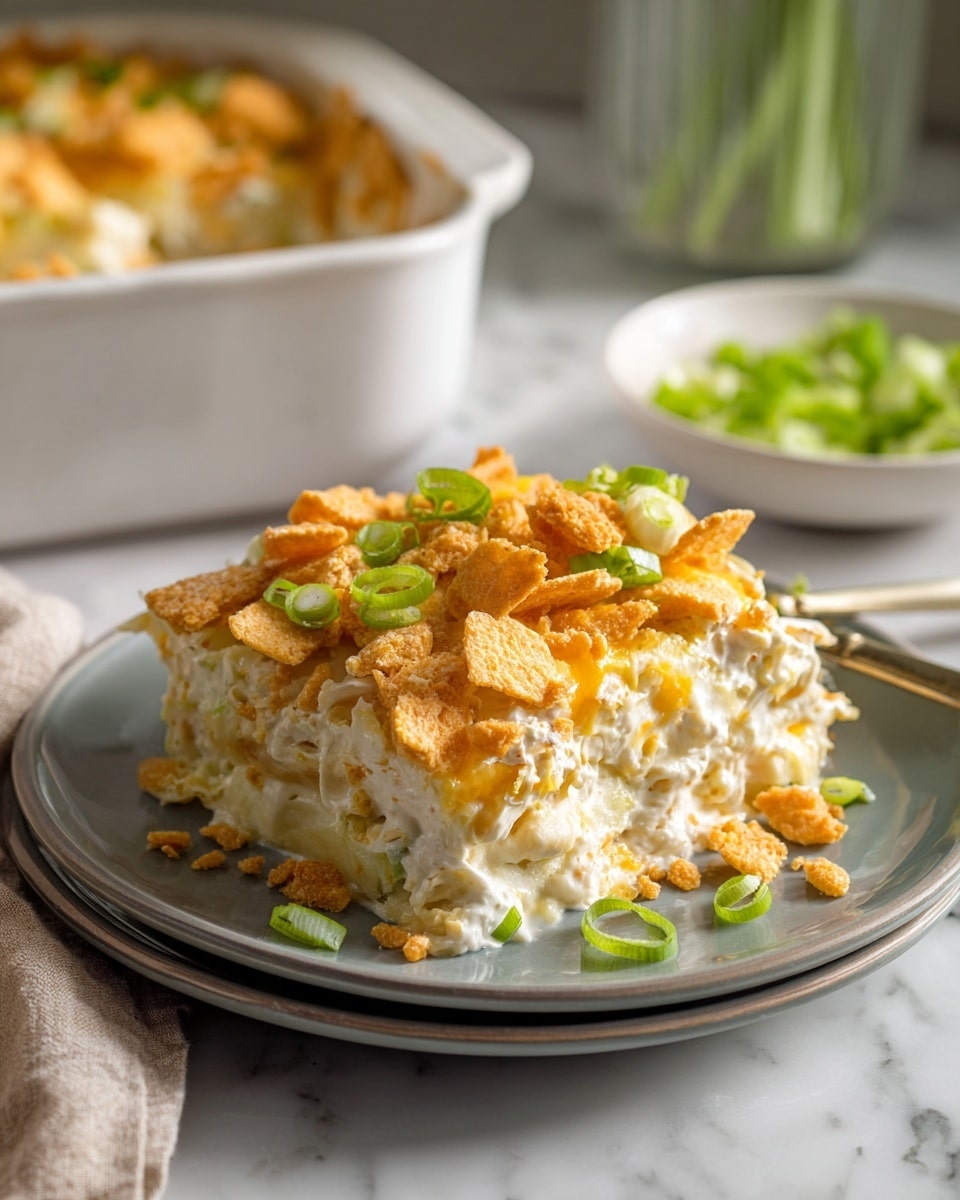 This image shows a close-up of a creamy layered casserole served on a gray plate on a white marbled surface. The dish has about four main layers: a creamy white sauce mixed with shredded pieces, topped with crumbled golden-brown crackers and small bits of orange sauce drizzled on top. Scattered over the dish are slices of green onions adding a fresh green color. In the background, there is a white casserole dish filled with the same layered food, and a small white bowl holding more sliced green onions. The whole scene has soft lighting and looks warm and inviting. photo taken with an iphone --ar 4:5 --v 7