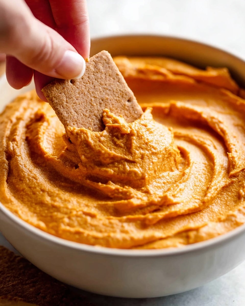 A white bowl filled with light brown pumpkin dip that has a creamy and smooth texture with swirls on the top. On one side of the dip, there is a swirl of white whipped cream sprinkled with a bit of brown spice. The bowl sits on a round wooden board surrounded by light green apple slices and rectangular light beige crackers laid out in a fan shape. A metal spoon rests on the wooden board next to the bowl. The setting is on a white marbled surface. photo taken with an iphone --ar 4:5 --v 7