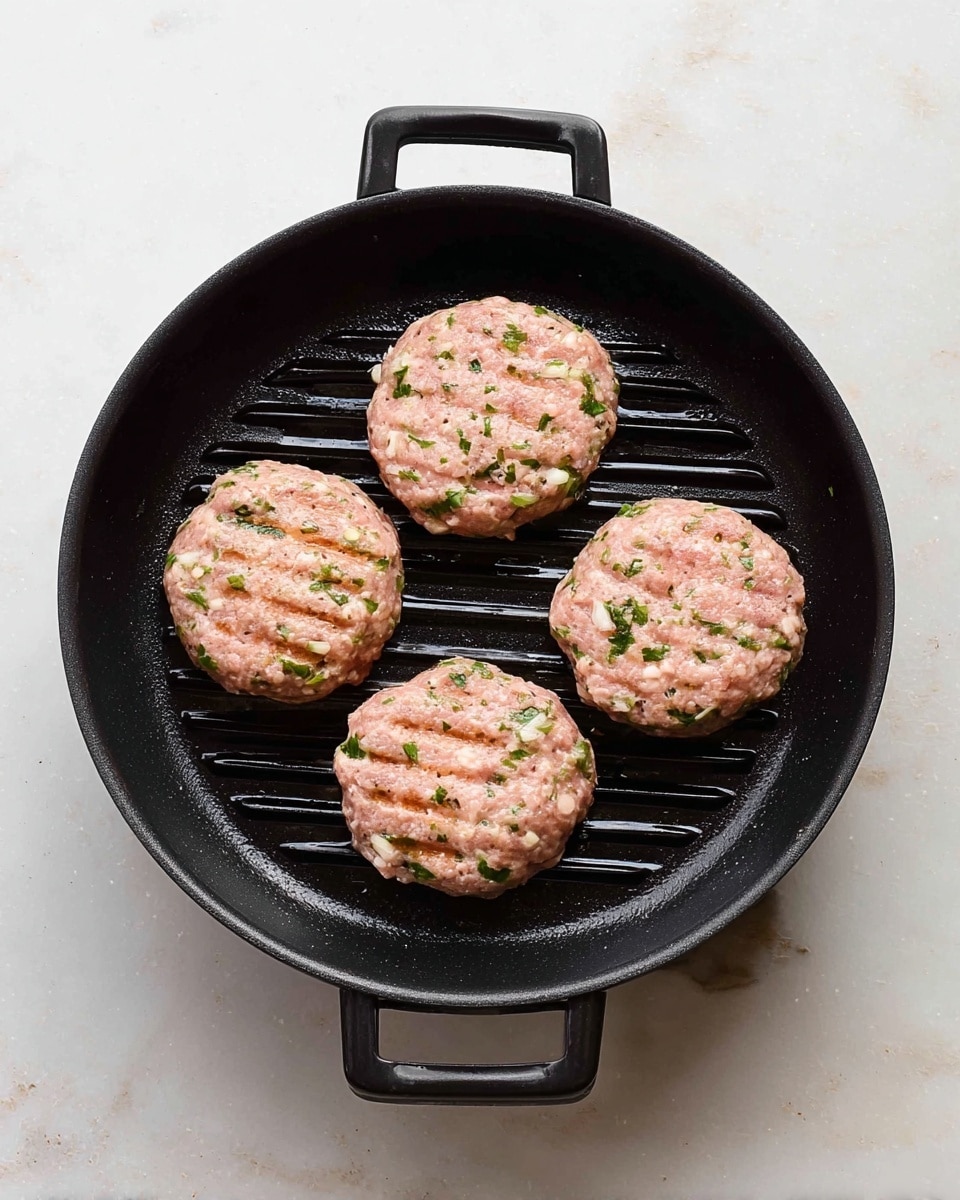 A black round grill pan holds four raw, round patties made from ground meat mixed with green herbs and small white pieces, evenly spaced on the pan's ridged surface. The patties are light pink with specks of green and white, showing a soft, textured appearance. The pan is set on a white marbled surface, with the handles visible on both sides. Photo taken with an iphone --ar 4:5 --v 7