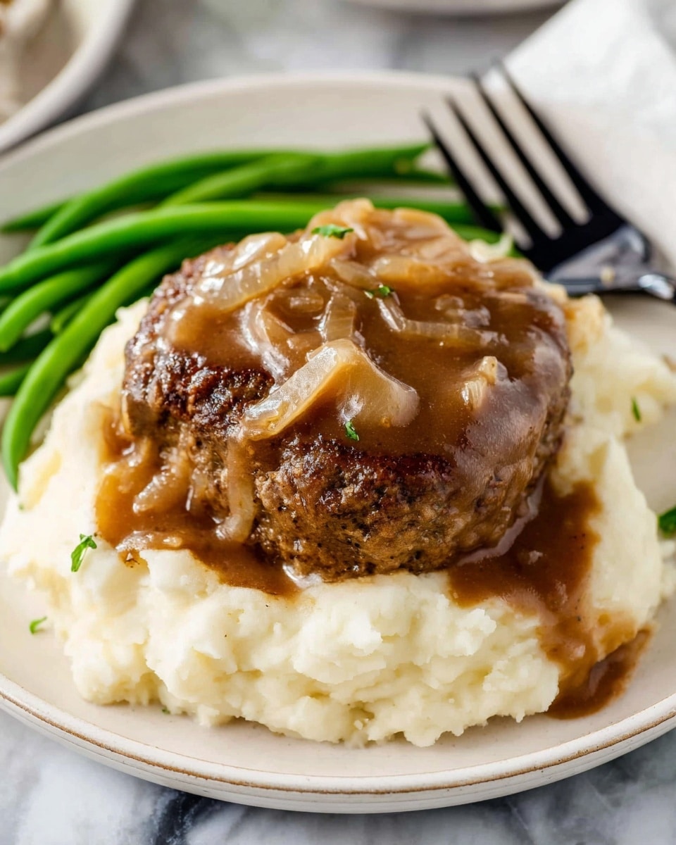 A white plate holds a layer of smooth, creamy mashed potatoes at the bottom, pale off-white in color with a soft texture. On top of the mashed potatoes lies a thick, browned meat patty with a slightly rough surface texture. Covering the meat patty is a generous layer of brown gravy with visible small chunks of cooked onions and a glossy finish. In the background, some green beans with a fresh, vibrant green color are placed near the top edge of the plate. The whole dish is set on a white marbled surface, with a black-handled fork positioned near the beans. photo taken with an iphone --ar 4:5 --v 7