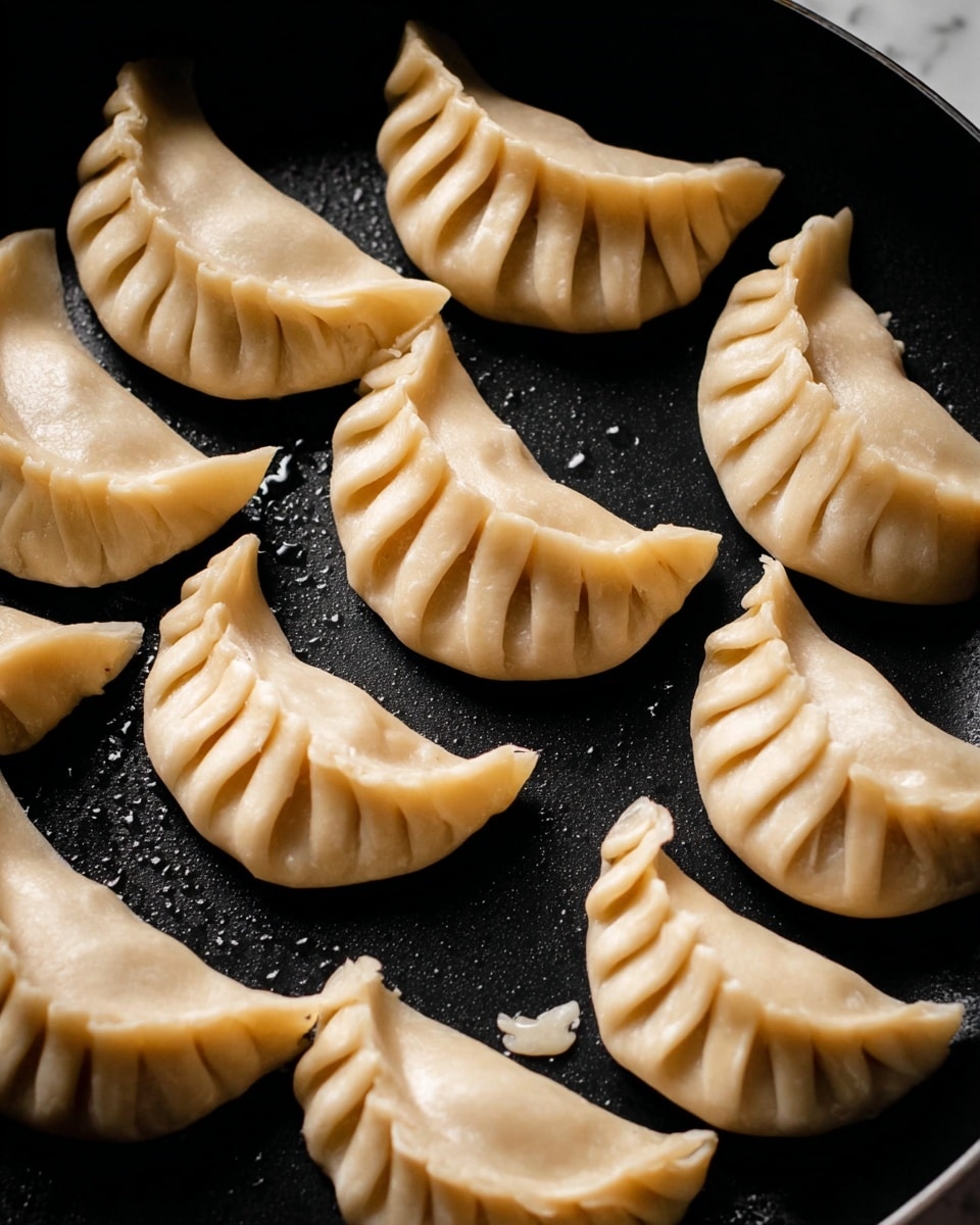 The image shows a close-up of twelve uncooked dumplings arranged in a black cooking pan with a few drops of oil shining under light. Each dumpling is folded in a crescent shape with neat pleats along the edges. Their dough is pale beige, smooth, and slightly translucent. The dumplings are placed in layers side by side, covering most of the pan surface. The background is a white marbled texture. Photo taken with an iphone --ar 4:5 --v 7