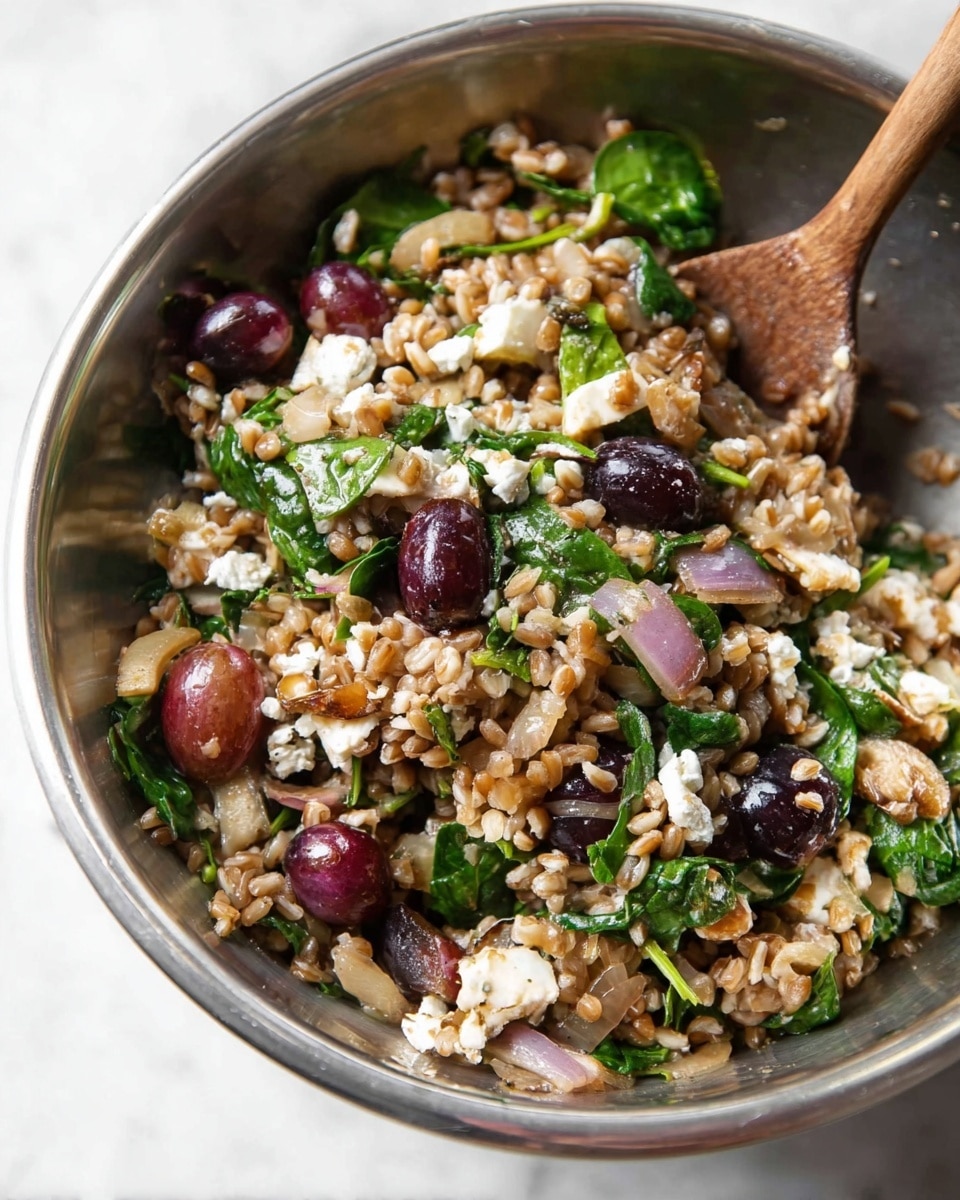 A close-up image of a metal bowl with a mixed salad inside, showing layers of light brown grains, halved dark red grapes, small white crumbly cheese pieces, bright green leafy spinach, and bits of caramelized onions, all stirred together with a wooden spoon on the right side of the bowl; the bowl is placed on a white marbled textured surface. Photo taken with an iphone --ar 4:5 --v 7