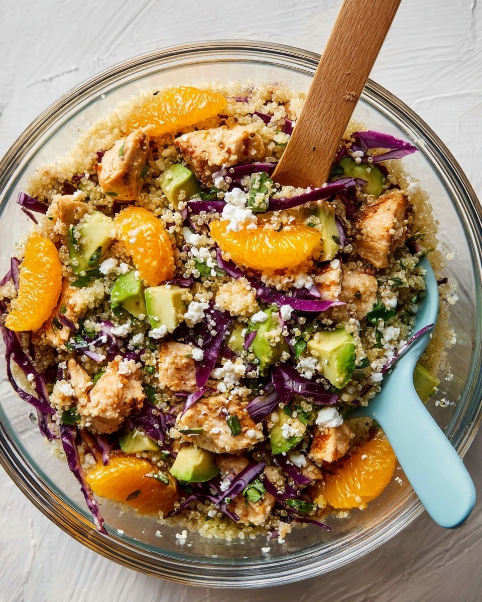 A clear glass bowl filled with a colorful salad made of four main layers: the base layer is light beige quinoa with a soft, grainy texture; scattered on top are bright orange segments with a juicy, slightly translucent look; small chunks of cooked light brown chicken pieces add a tender, slightly browned texture; and fresh green avocado cubes with a smooth, creamy surface. Thin, dark purple shredded cabbage pieces are spread throughout, adding a crisp look, along with small bits of white crumbled cheese and finely chopped green herbs giving a fresh touch. A light blue silicone spatula with a wooden handle is resting inside the bowl on a white marbled surface. Photo taken with an iphone --ar 4:5 --v 7