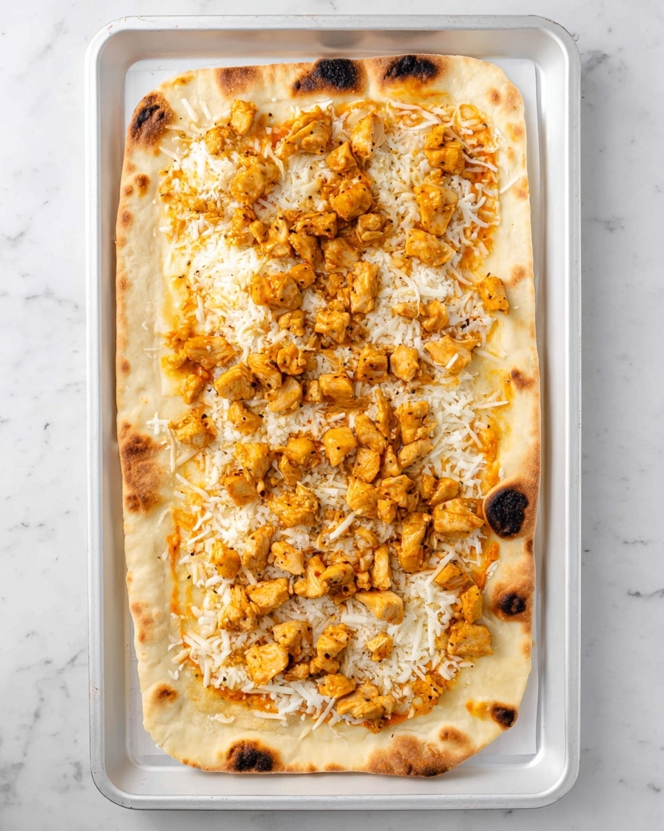 A rectangular flatbread with light golden brown spots and slightly charred edges is placed in the center of a white baking pan. The bottom layer is a thin spread of orange sauce, followed by a layer of white shredded cheese scattered evenly across the bread. On top, there are small chunks of cooked chicken colored golden yellow-orange with some slight seasoning visible. The background is a white marbled surface. photo taken with an iphone --ar 4:5 --v 7