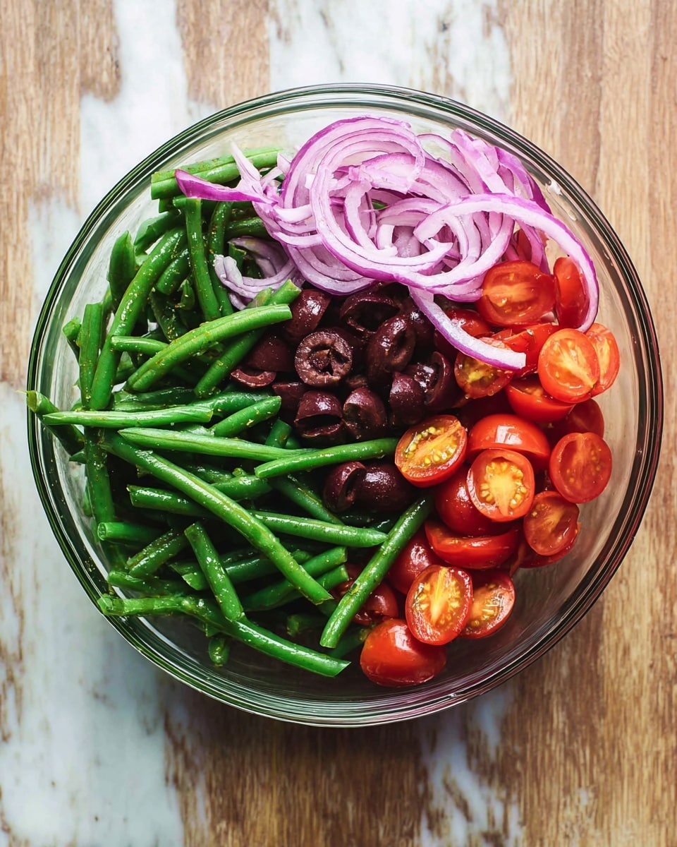 A clear glass bowl sitting on a white marbled surface holds a colorful salad with four distinct layers. The bottom layer is bright green beans with a shiny texture, topped with halved cherry tomatoes showing their juicy red interiors in the middle left section. To the upper left, thin slices of light purple onion are placed, while dark purple, halved small olives form the upper middle layer. photo taken with an iphone --ar 4:5 --v 7
