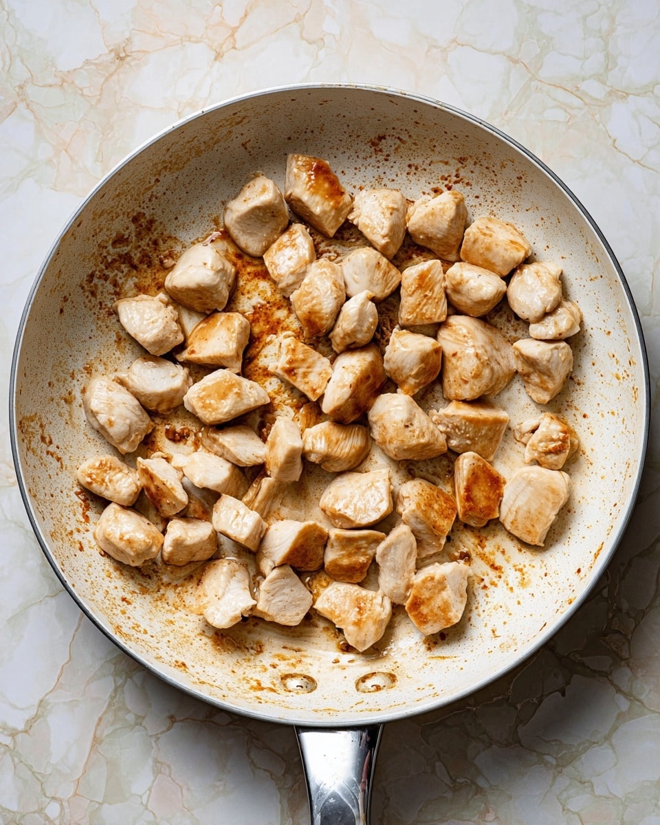 A white pan holds many small, cooked pieces of chicken scattered unevenly inside. Each piece shows a light golden brown color on some sides, with smooth and slightly shiny white parts. The pan's surface has brown, uneven marks and spots from cooking, especially around the edges. The background features a white marbled texture surface under the pan's silver handle. photo taken with an iphone --ar 4:5 --v 7