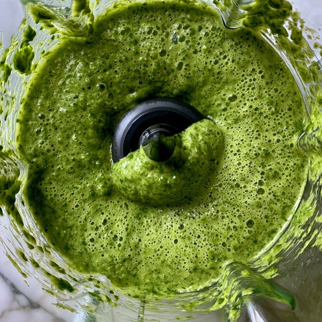 This image shows a close-up of a blender bowl with green sauce inside it. The sauce is thick and bubbly with small bits and a smooth oily texture. The blender blade is black and placed right in the center, partly covered by the green sauce. The sides of the blender bowl are stained with green sauce splashes. The surface under the blender has a white marbled look. Photo taken with an iphone --ar 4:5 --v 7