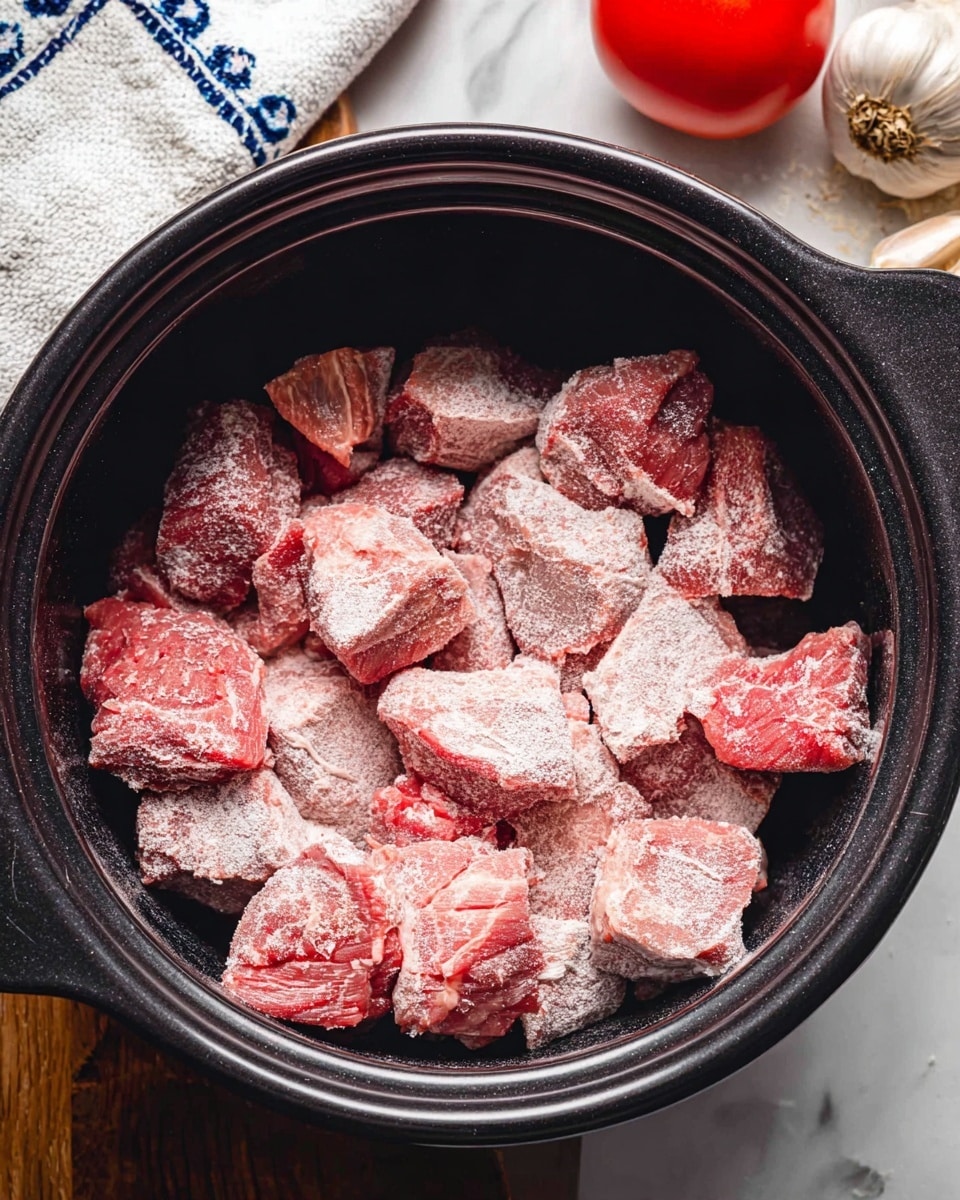 Chunks of raw red meat coated in a light layer of white flour fill a black pot. The meat pieces vary in size and shape, showing rough textures and some cracks on their surfaces. To the upper left of the pot, there is a segment of a white cloth with blue patterns, a whole bulb of garlic with brown tips, and a plump red tomato, all set on a white marbled surface. Photo taken with an iphone --ar 4:5 --v 7