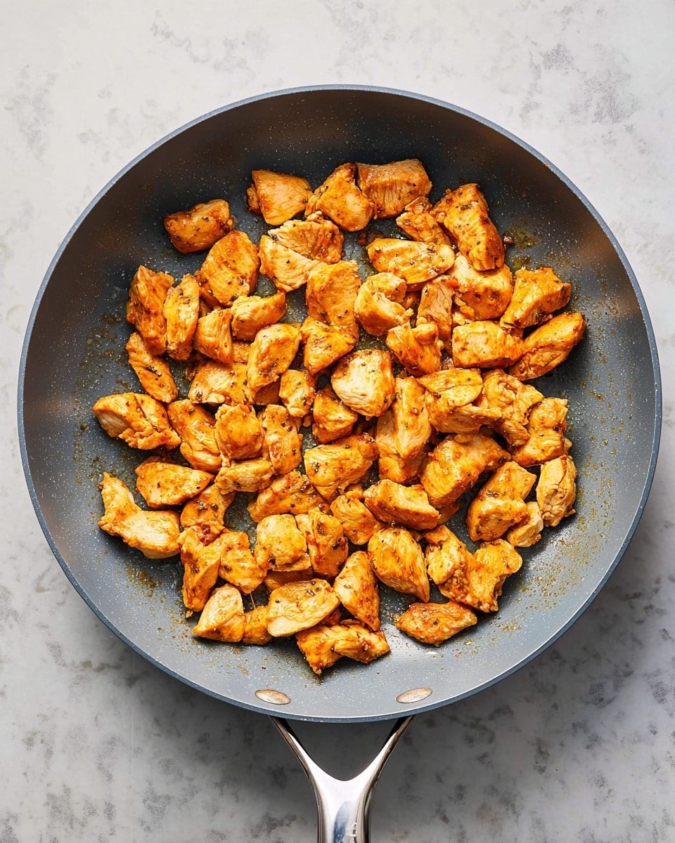 A gray pan filled with one layer of small pieces of cooked chicken, each piece golden brown with a slightly crispy texture on the edges. The chicken pieces are placed evenly across the pan's surface, showing a rich mix of seared orange and light brown shades. The pan handle is shiny metal, resting on the side, and the pan sits on a white marbled textured surface. photo taken with an iphone --ar 4:5 --v 7