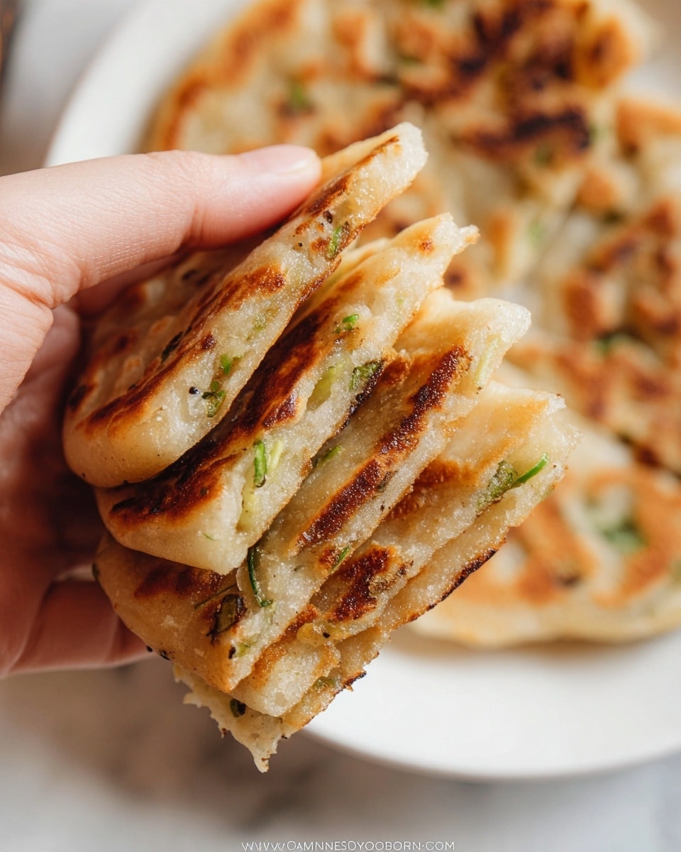A woman's hand is holding three thin layers of golden brown scallion pancakes stacked together, showing their crispy, slightly charred edges and flaky texture. Each pancake layer has a light, slightly translucent dough with visible bits of green scallions scattered inside. The background shows more scallion pancakes resting on a white plate, placed on a white marbled surface. The overall look is warm and inviting with a close-up focus on the texture and layers of the pancakes photo taken with an iphone --ar 4:5 --v 7
