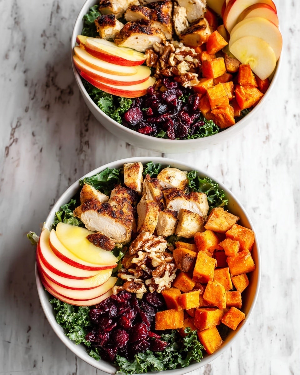 Two white bowls filled with layered salad sit on a white marbled surface. Each bowl has a base layer of dark green leafy kale, topped with grilled chicken pieces with a slight char and golden color on the top left side. Below the chicken are small burnt orange cubes of roasted sweet potatoes. On the right side of the bowls, there are thin, even slices of red and yellow apple arranged in a fan shape. In the middle, between apple and chicken, there are chopped nuts that look light brown and dried, dark red cranberries scattered next to the nuts and sweet potatoes. The overall look is colorful, fresh, and neatly arranged. Photo taken with an iphone --ar 4:5 --v 7
