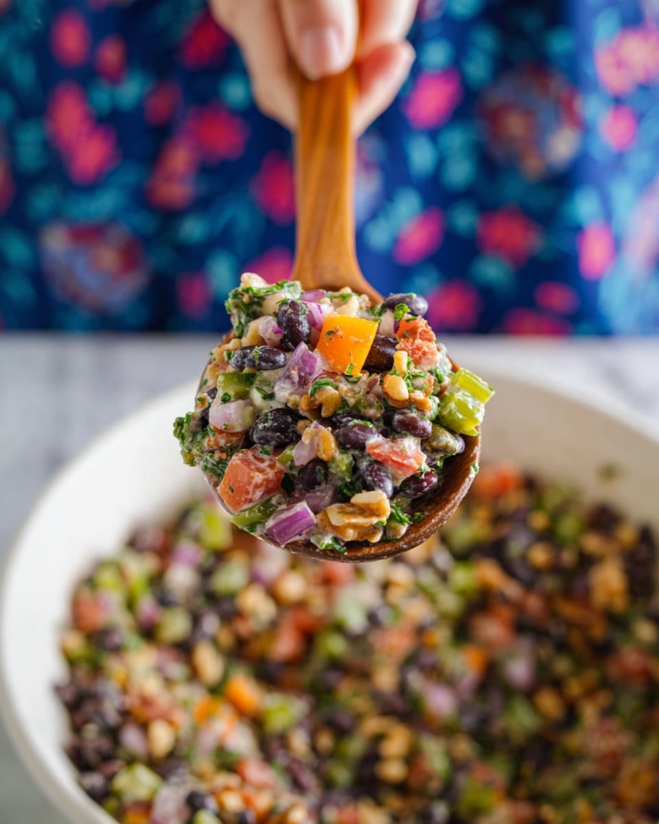 A wooden spoon held by a woman's hand with a mix of colorful salad ingredients is shown close up. The salad has many layers with black beans, green herbs, diced orange tomatoes, red onions, and chopped nuts all mixed together creating a rich texture with bits of cream-colored dressing. This mix sits above a large white bowl filled with the same ingredients. The background is a blue fabric with a floral pattern, and the whole scene is set on a white marbled surface. photo taken with an iphone --ar 4:5 --v 7