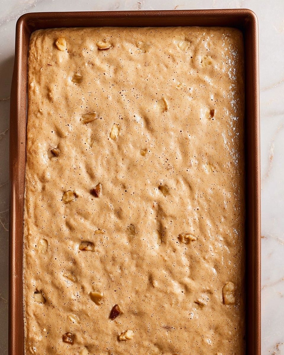 A close-up view of a rectangular brown baking tray filled with a thick, light brown batter with small, unevenly spread pieces of fruit embedded inside. The batter has a slightly bubbled and textured surface with varied tiny holes and a puffy appearance. The background is a white marbled surface. photo taken with an iphone --ar 4:5 --v 7