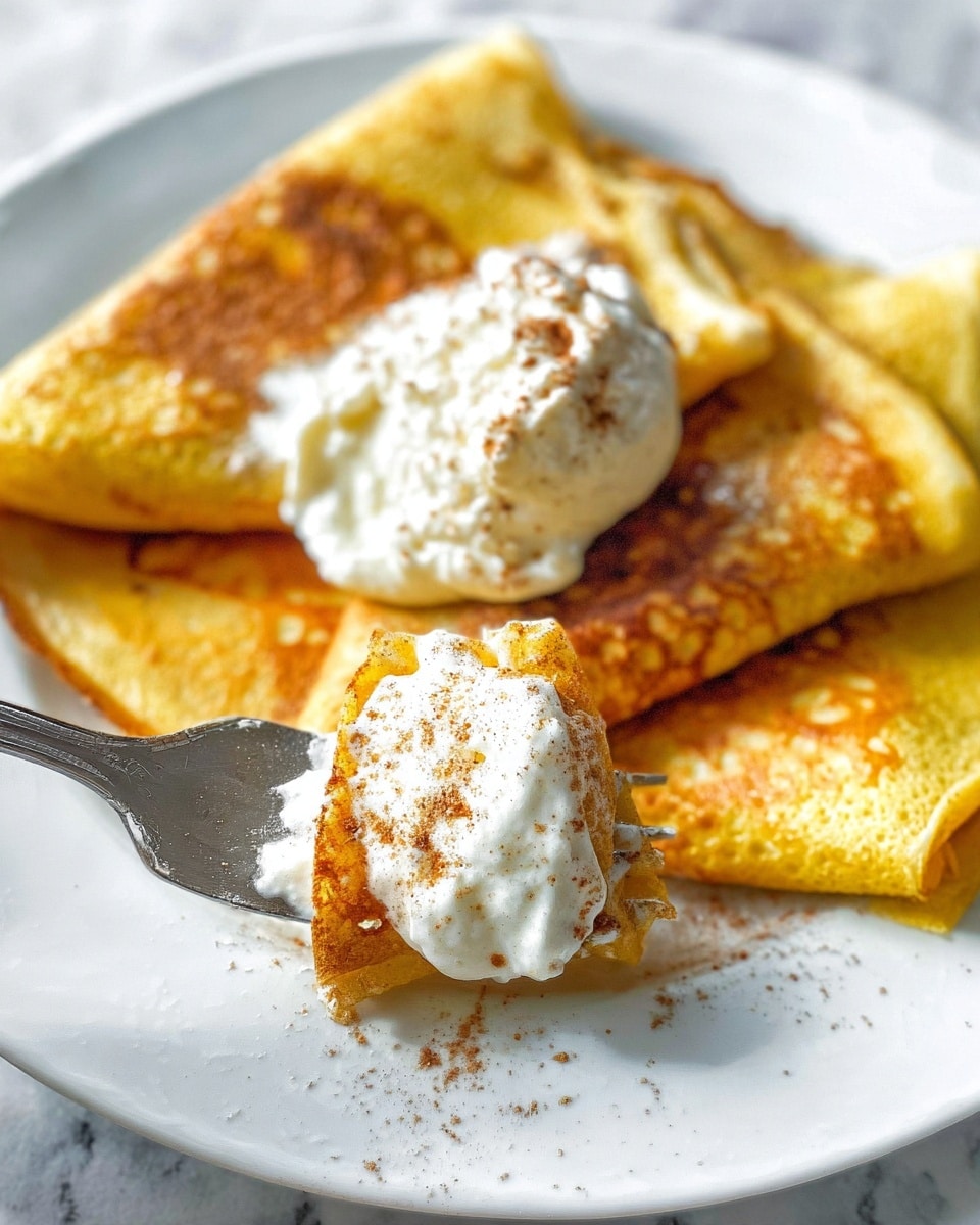The image shows three folded golden-yellow crepes placed side by side on a white plate with a white marbled surface underneath. On top of the crepes, there is a small dollop of white cottage cheese sprinkled lightly with brown cinnamon powder. In the foreground, there is a silver fork holding up a bite of crepe layered with a thick, fluffy white cream topped with a light sprinkling of cinnamon, creating a soft texture contrast. The lighting highlights the warm colors of the crepes and the creamy whiteness of the cheese and topping, making the dish look inviting and fresh. Photo taken with an iphone --ar 4:5 --v 7